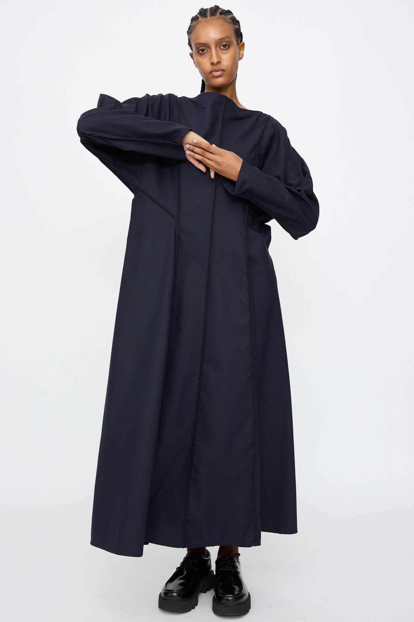 A person stands against a plain background wearing the Baserange Nika Dress in Dama Navy, a long sleeve silk wool blend maxi with a boat neck, paired with black shoes and arms positioned asymmetrically.