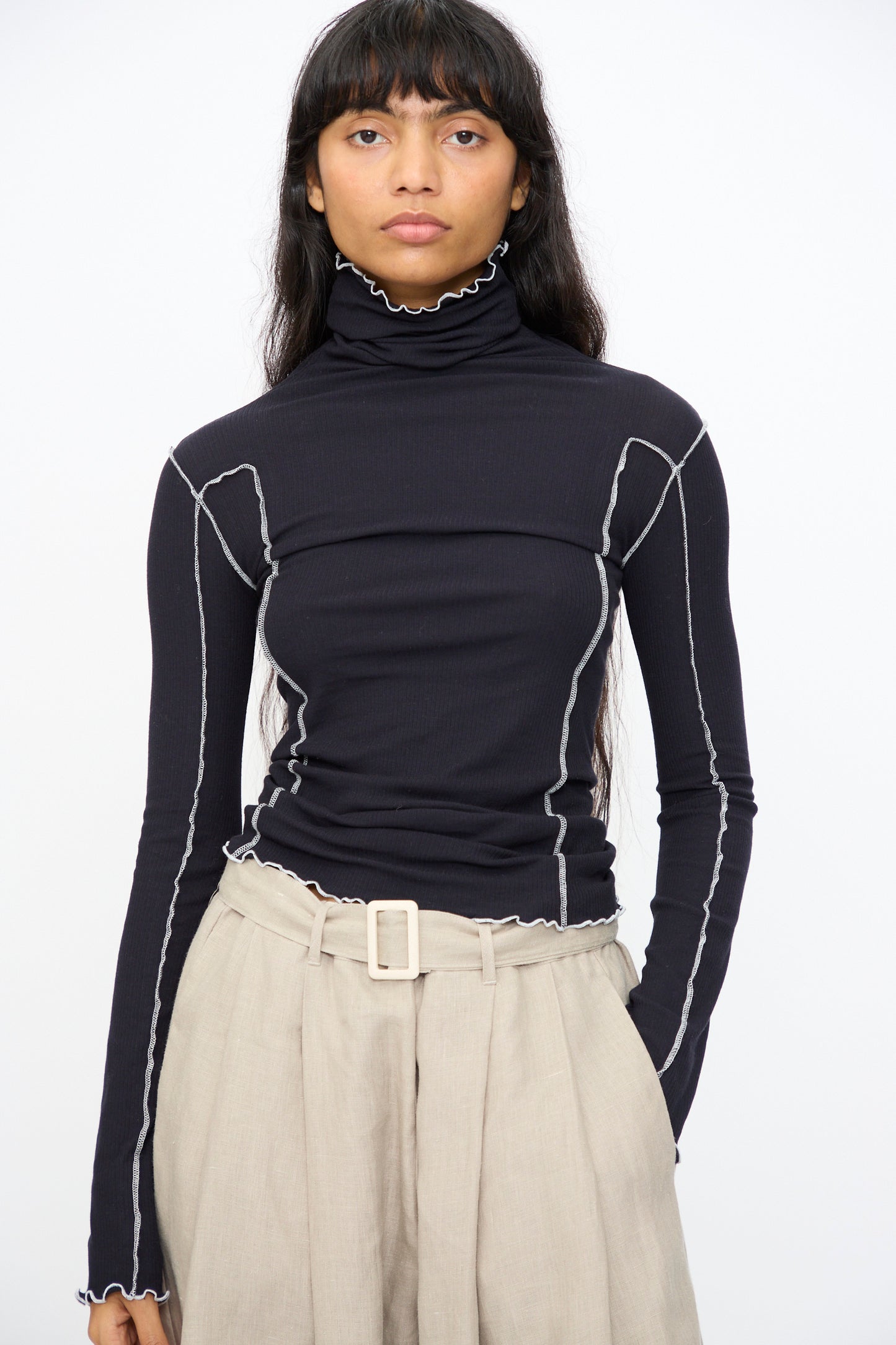 A person wears the Baserange Omato Turtleneck in Murano Navy and Undyed, featuring contrast exposed seams, paired with beige high-waisted pants, standing against a plain white background.