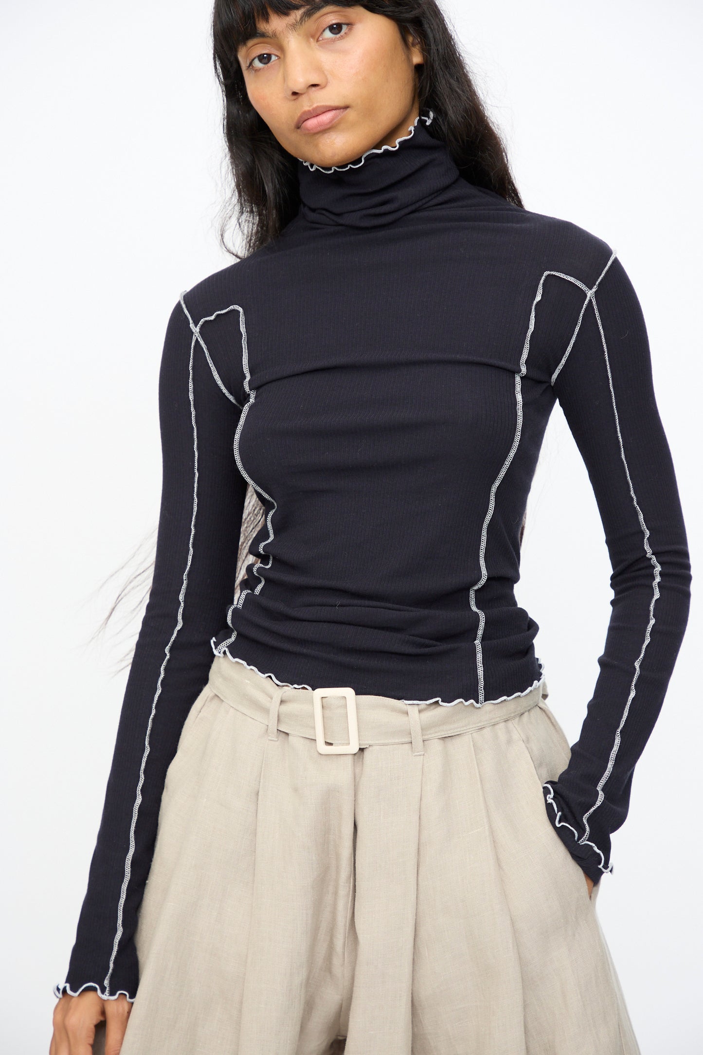 A person wears the Baserange Omato Turtleneck in Murano Navy and Undyed, featuring contrast exposed seams, paired with beige high-waisted pleated trousers and a belt, standing against a plain white background.