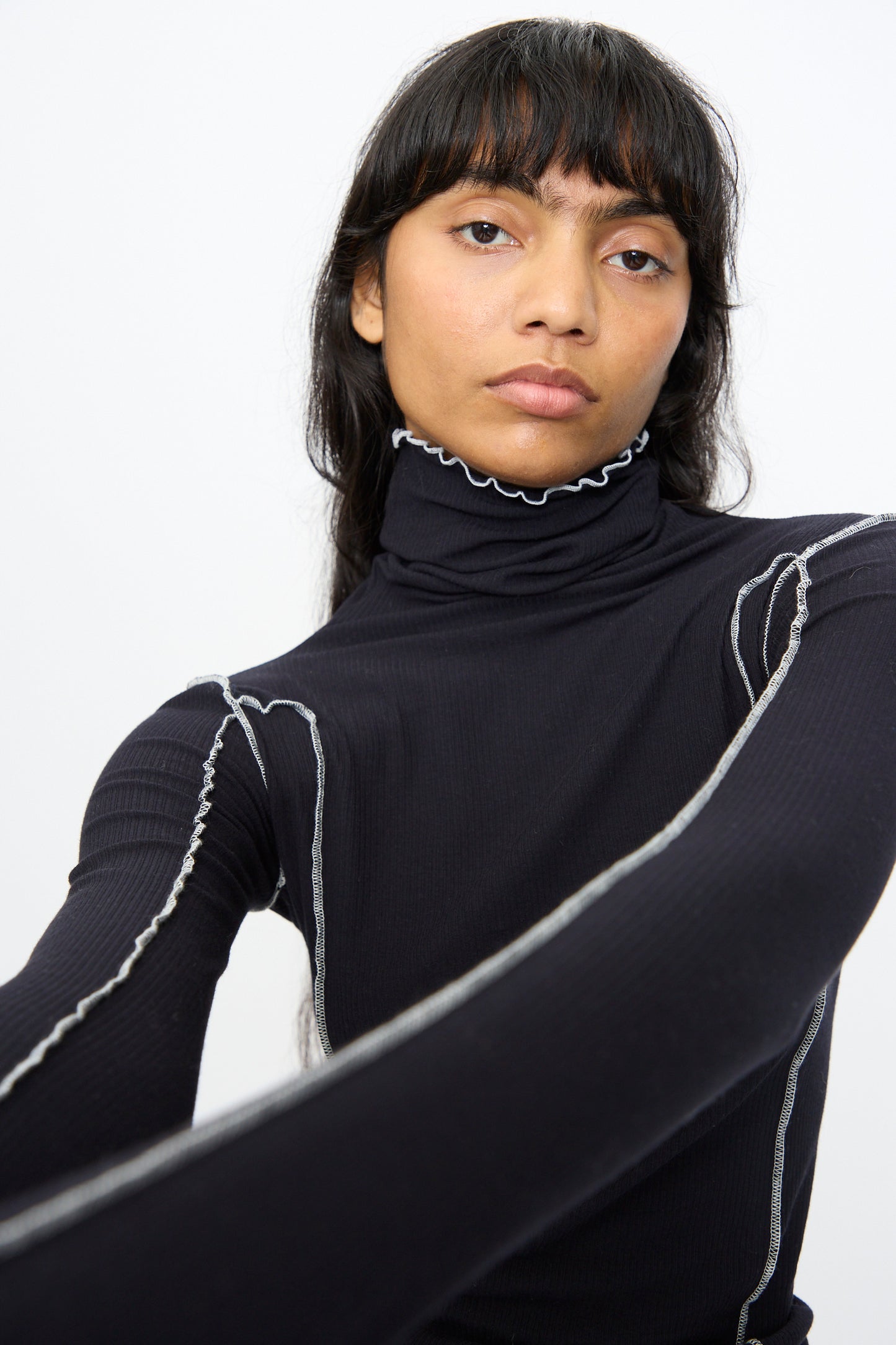 A person with long dark hair and bangs wears the Baserange Omato Turtleneck in Murano Navy and Undyed, featuring ribbed organic cotton with white stitching, looking at the camera against a plain light background.
