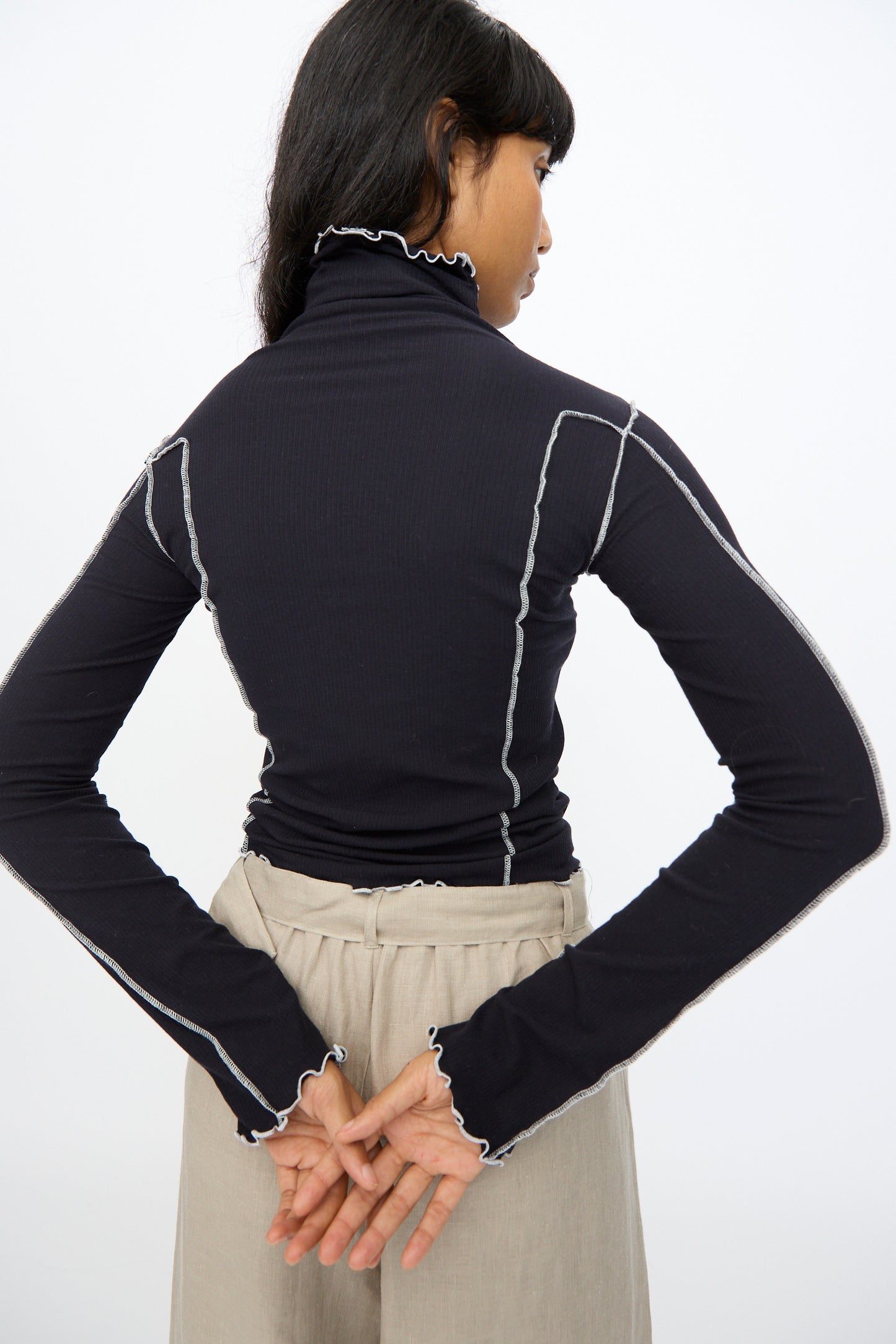 A woman stands with her back to the camera, wearing the Baserange Omato Turtleneck in Murano Navy and Undyed, her hands clasped behind her back.