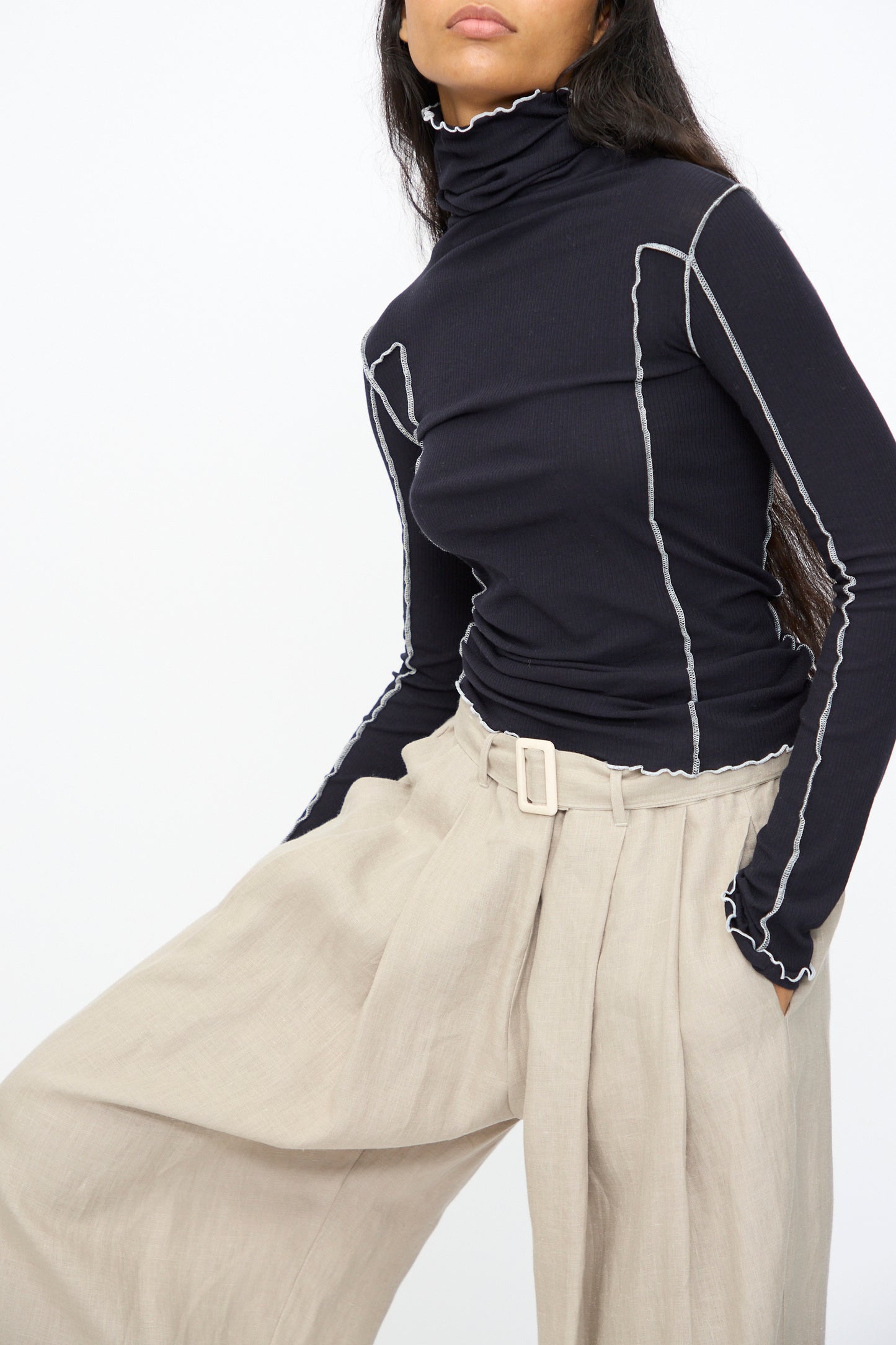 A person wears the Baserange Omato Turtleneck in Murano Navy and Undyed, featuring ribbed organic cotton and contrast exposed seams, paired with beige wide-leg belted pants, posing against a plain white background.