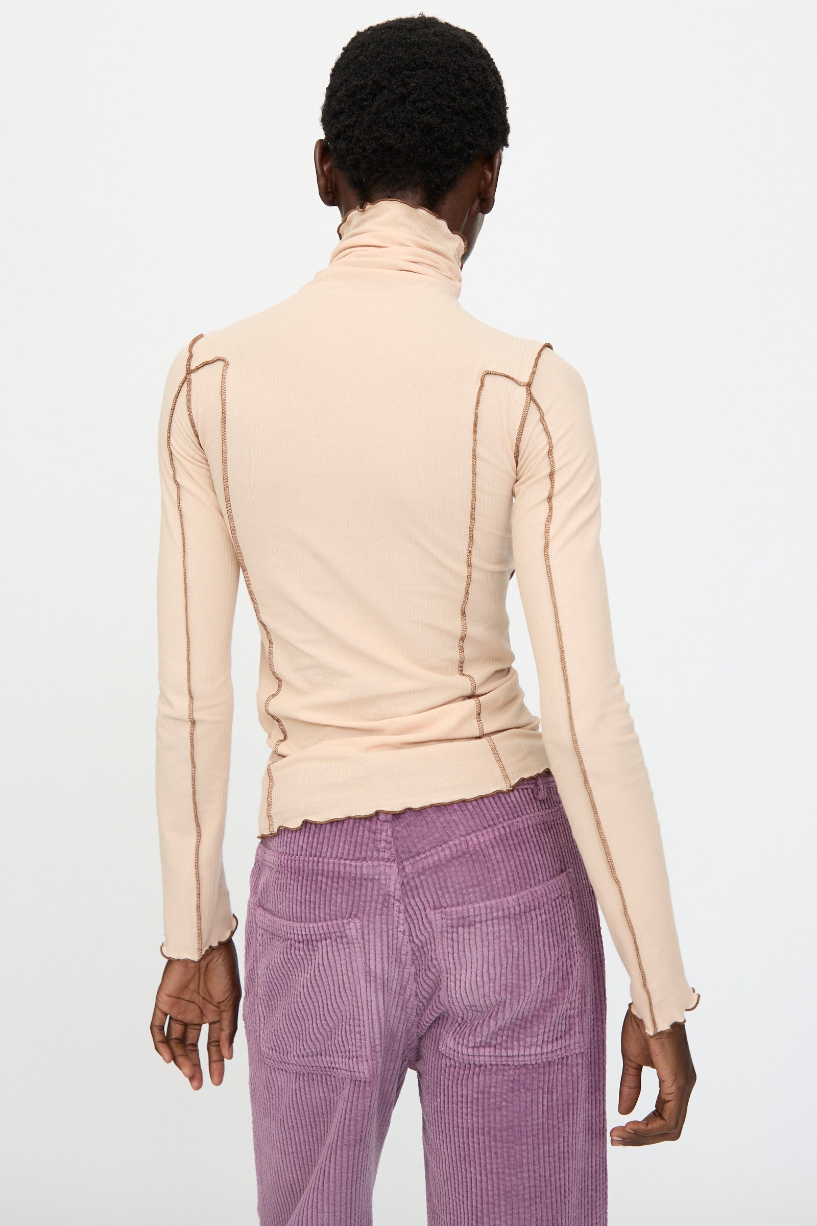 A person stands with their back to the camera, wearing the Baserange Omato Turtleneck in Tin Beige and Ocular, paired with high-waisted purple corduroy pants.