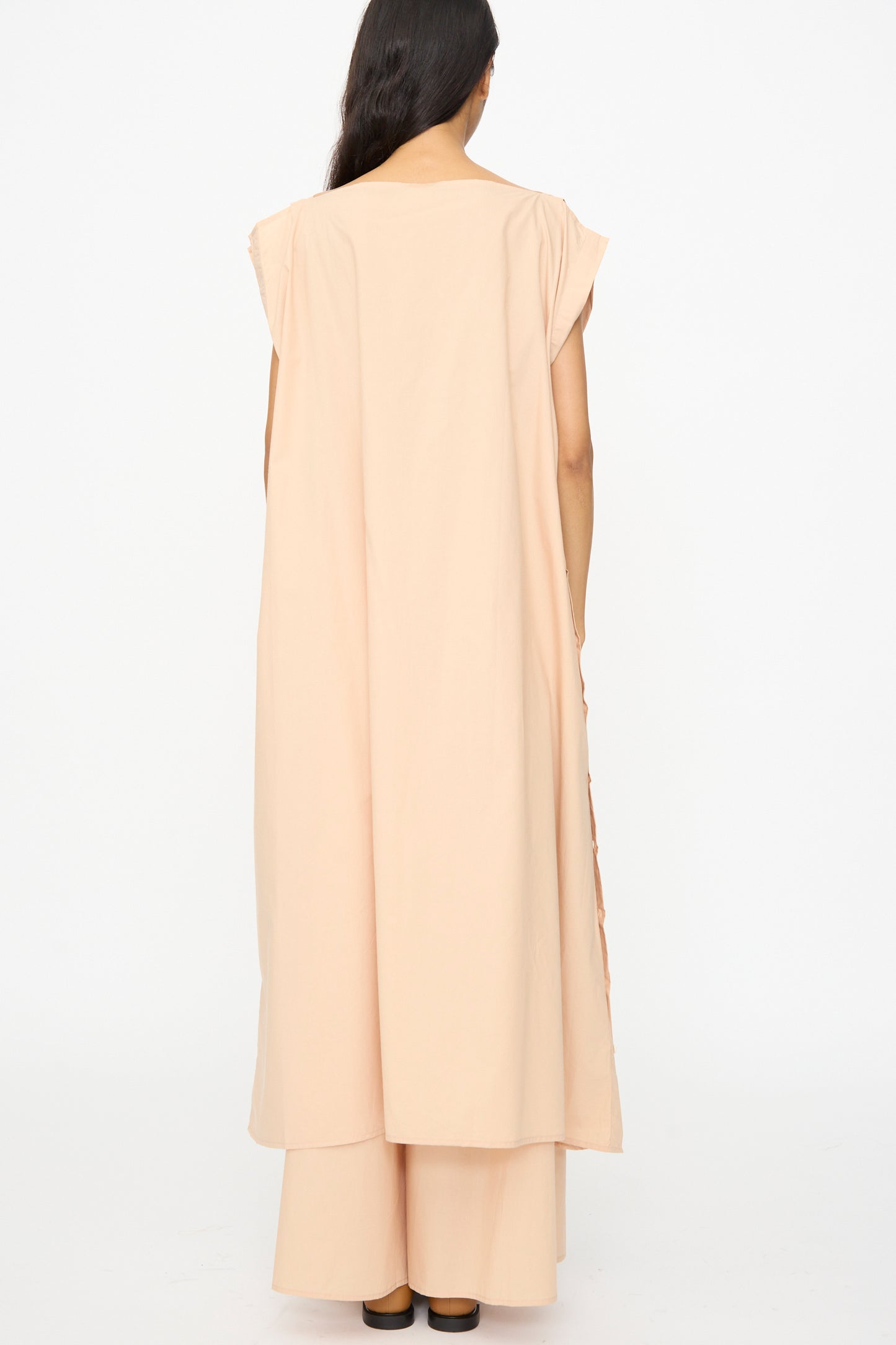 A person faces away, wearing the Baserange Part Dress in Andradita Sand over wide-legged pants and black shoes, standing against a plain white background.