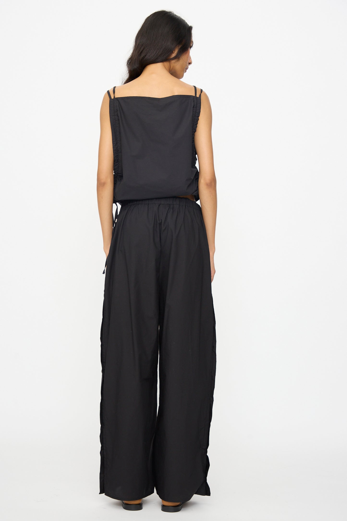 A person with long dark hair faces away, wearing a sleeveless black top and Baserange’s Part Pants in Black—loose cotton poplin wide leg pants with an elastic waist—against a plain white background.