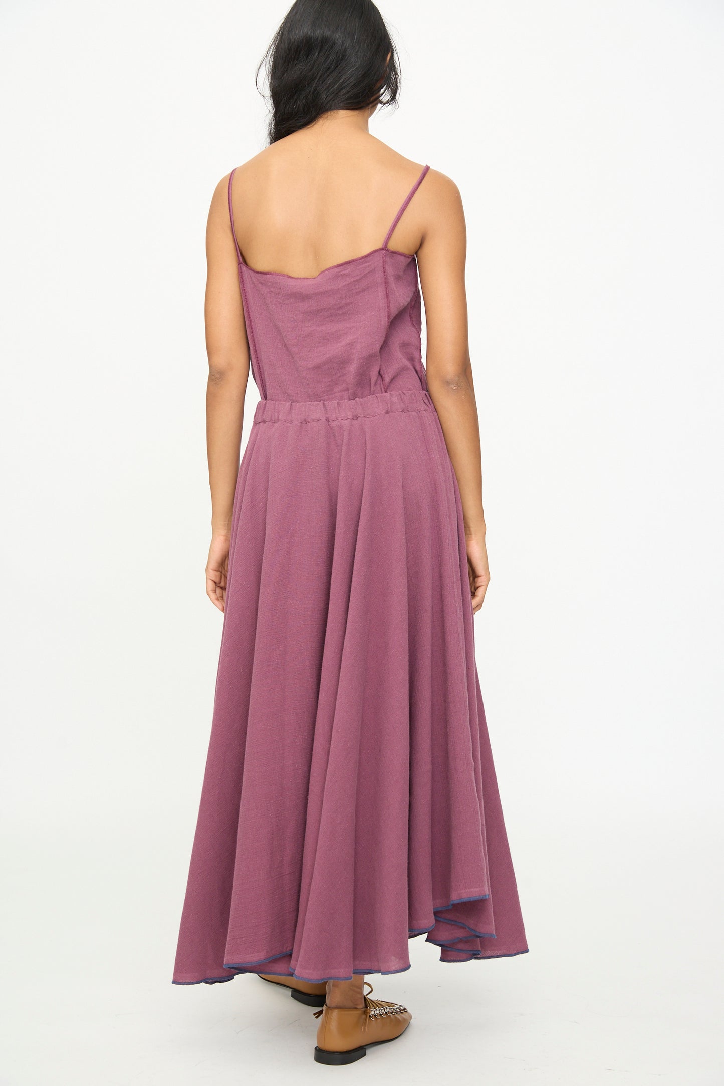 A person stands with their back to the camera, wearing the Baserange Rene Skirt in Aubergine—an ankle-length piece crafted from a linen cotton blend—paired with brown shoes, against a plain white background.