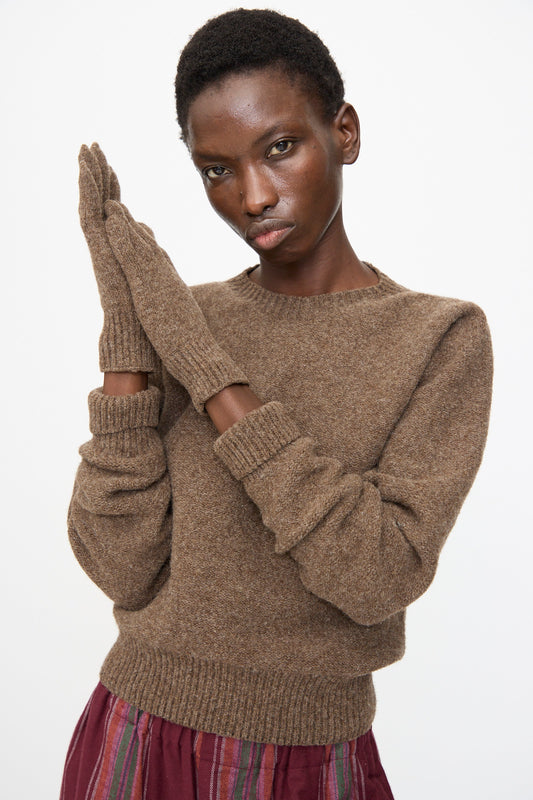 A person stands against a plain white background, hands pressed together, wearing Baserange Ribbed Gloves in Burel Brown along with a brown sweater and a maroon striped skirt.