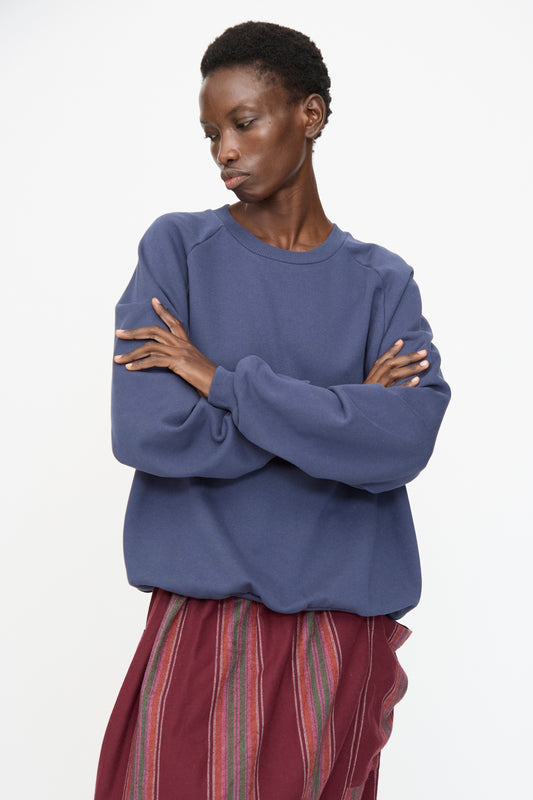 A person stands with arms crossed, wearing the Baserange Rim Raglan Sweatshirt in Echo Blue and a red vertically striped skirt, gazing to the side against a plain white background.