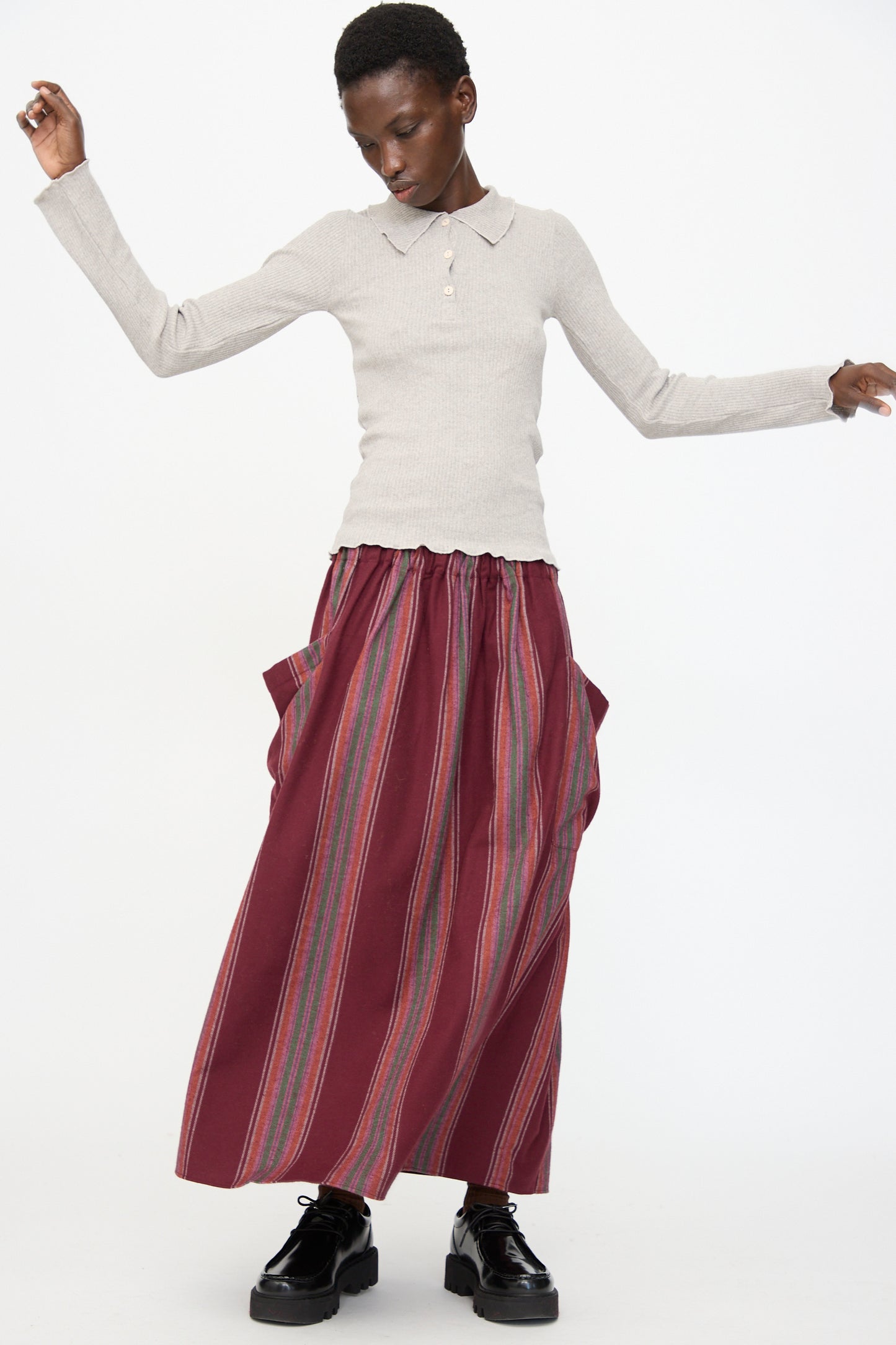 A person wearing a light gray long-sleeve shirt, black platform shoes, and the Baserange Snell Skirt in Maroon Stripe stands against a plain white background.