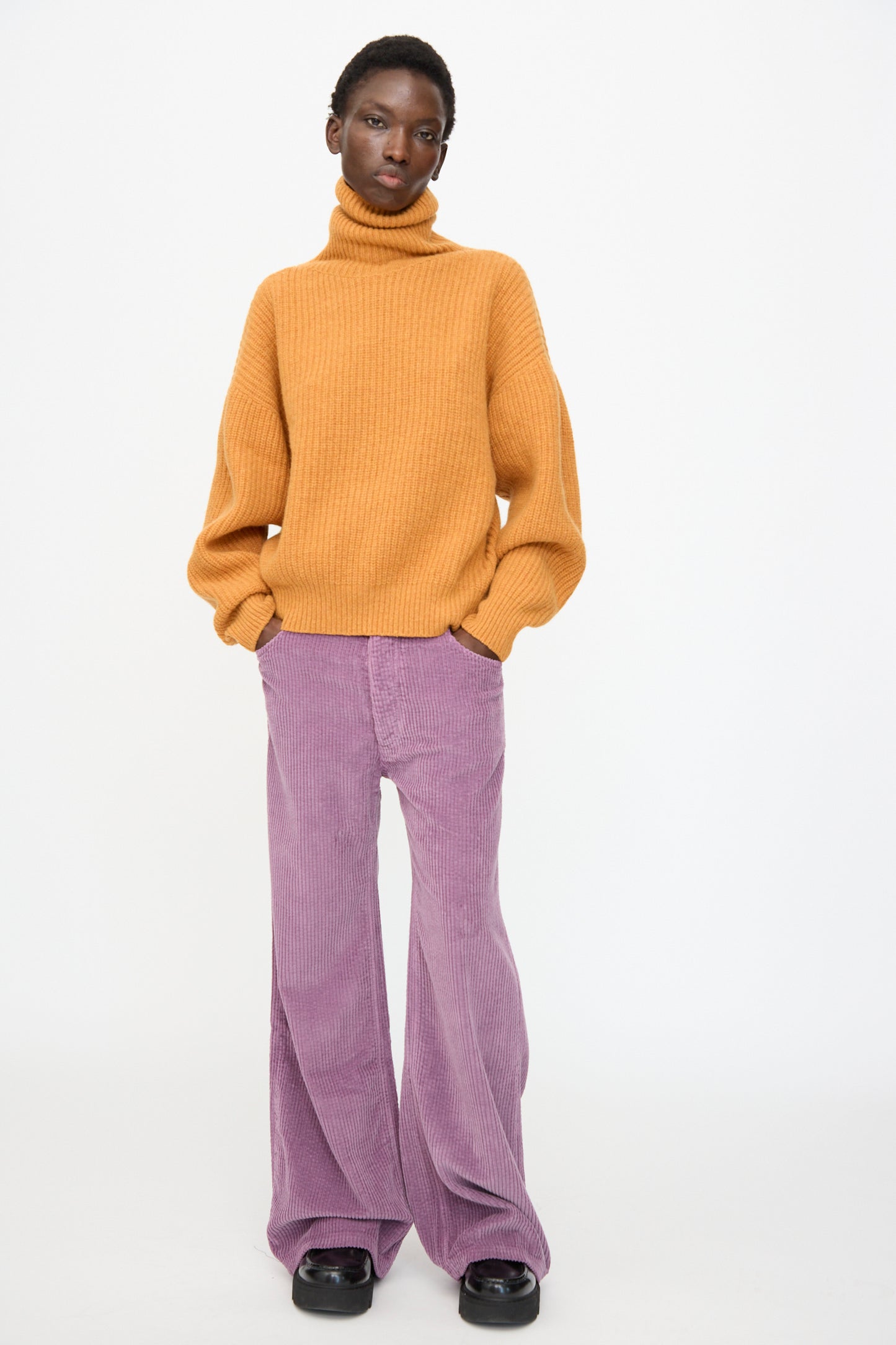 Person standing against a plain background, wearing an orange turtleneck sweater, Baserange Tajo Wide Pants in Cue Purple, and black shoes.