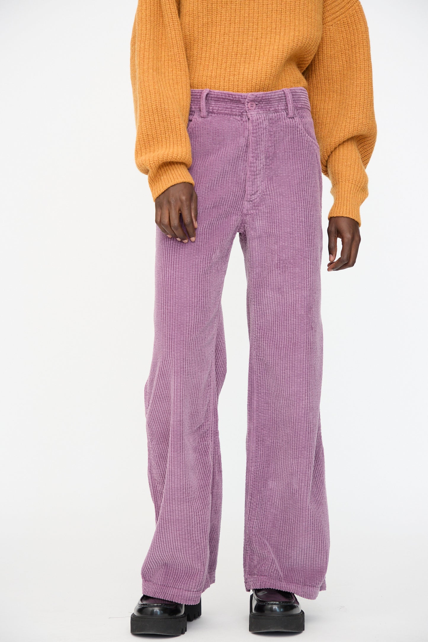 A person stands wearing a mustard yellow sweater and Baserange's Tajo Wide Pants in Cue Purple, paired with black shoes, against a plain background.