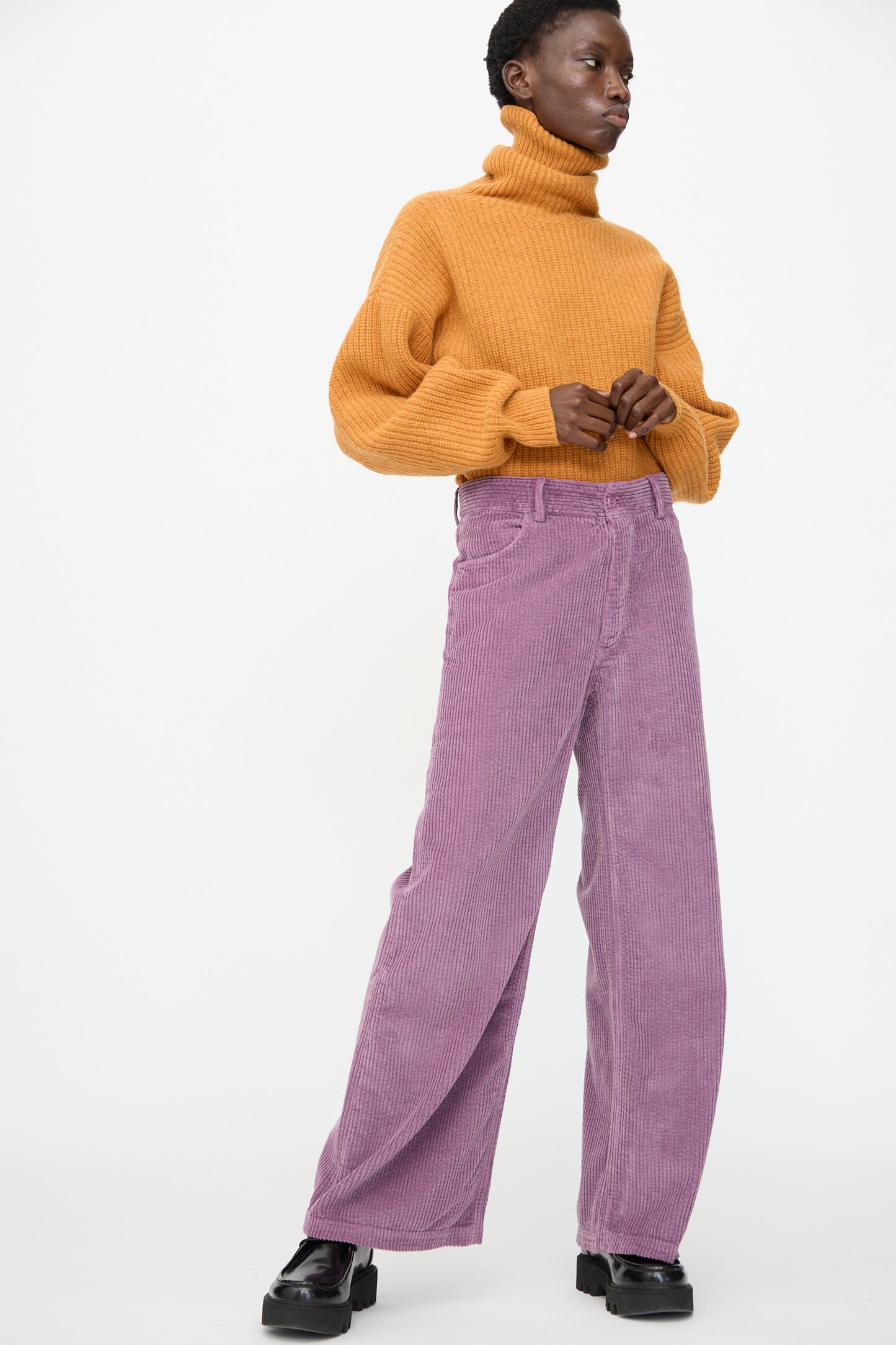 A person stands against a plain background, wearing a mustard turtleneck sweater, Baserange Tajo Wide Pants in Cue Purple, and black platform shoes.