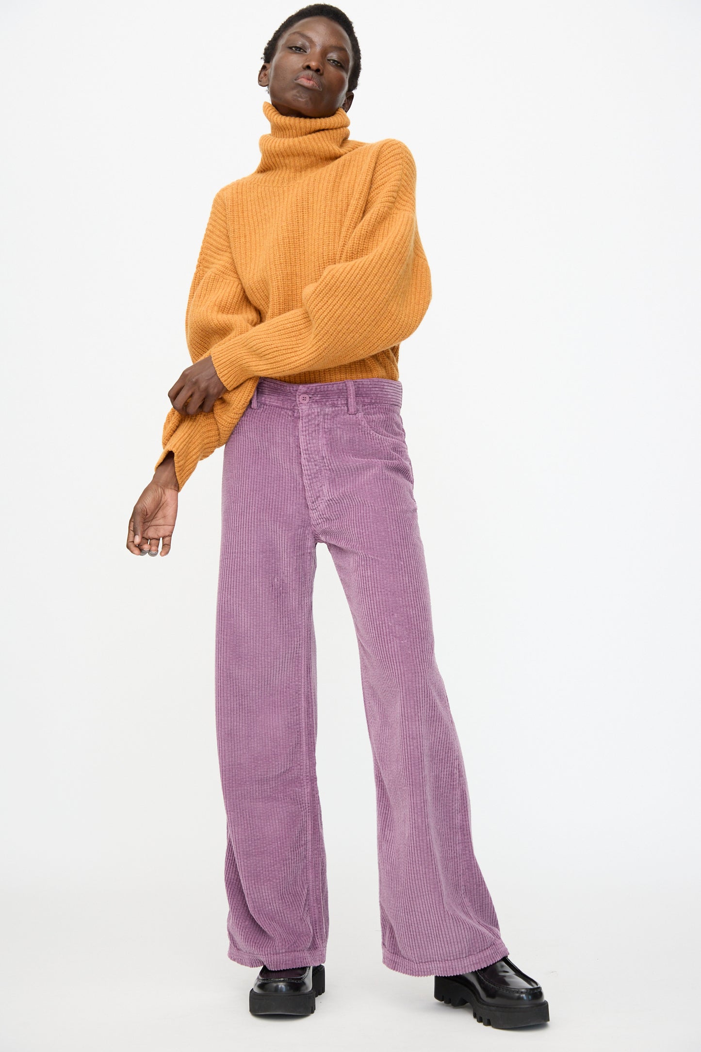 A person stands against a white background wearing a mustard turtleneck sweater, Baserange Tajo Wide Pants in Cue Purple, and black platform shoes.