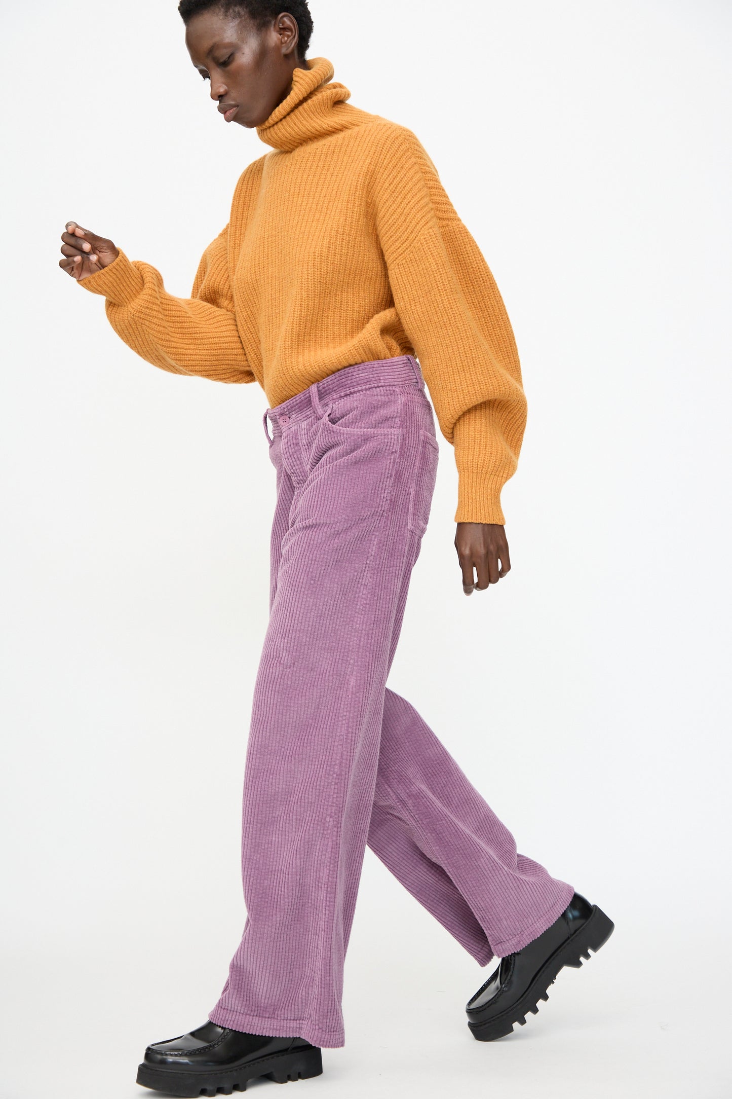 A person wearing a mustard yellow turtleneck, Baserange Tajo Wide Pants in Cue Purple, and black shoes walks against a white background.