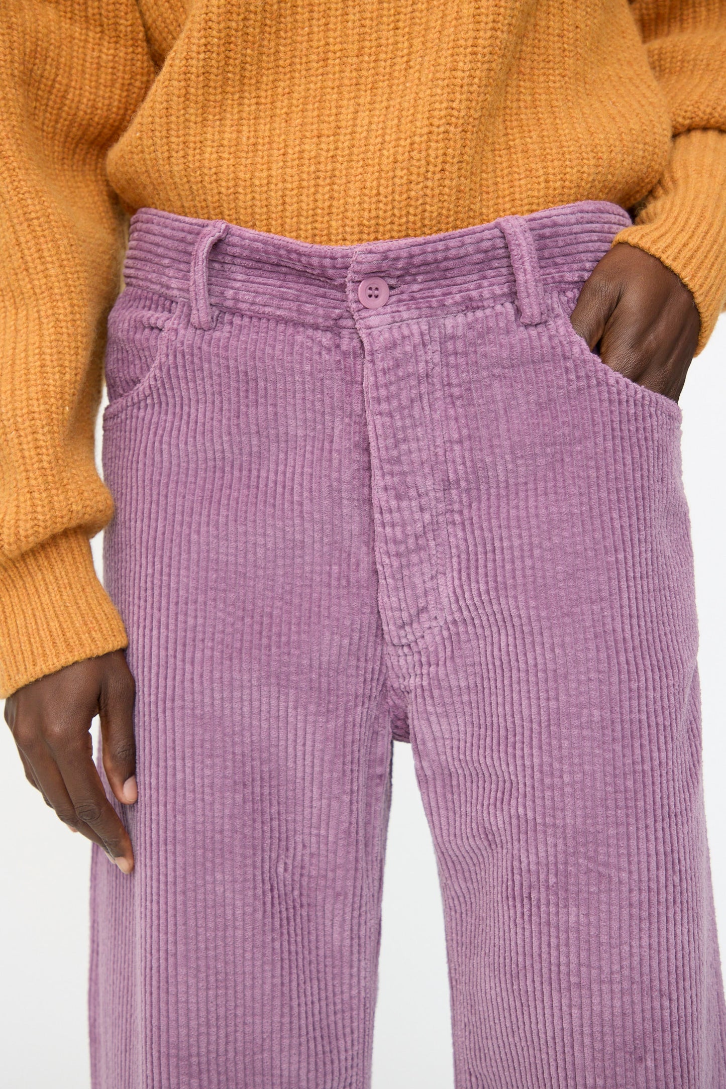 A person wearing a mustard yellow sweater and Baserange Tajo Wide Pants in Cue Purple—relaxed fit, high-waisted, made from organic cotton corduroy—with one hand in a pocket.