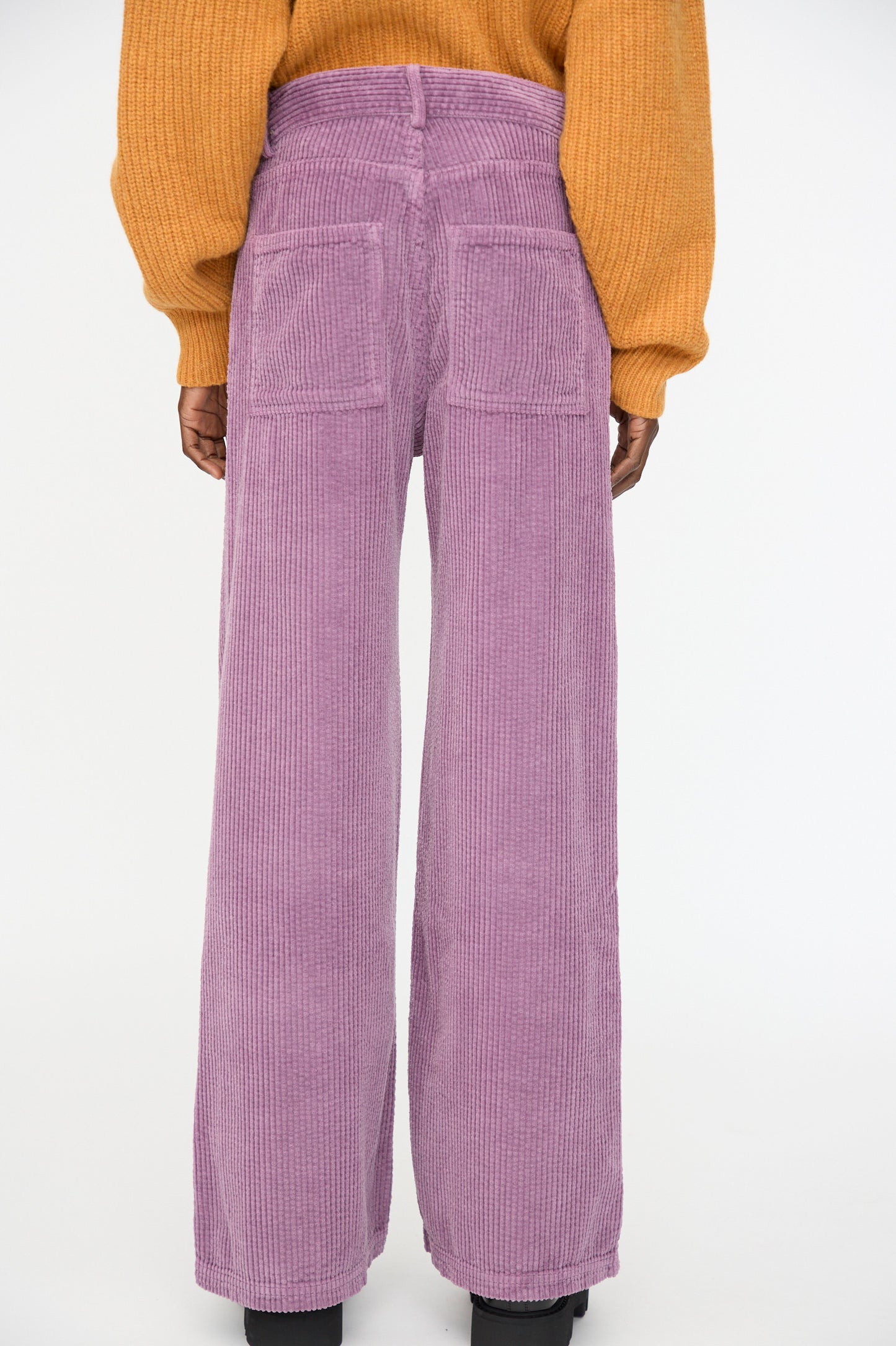 A person shown from the back wears a mustard yellow sweater and Baserange Tajo Wide Pants in Cue Purple—relaxed fit, high-waisted, and made from organic cotton corduroy.