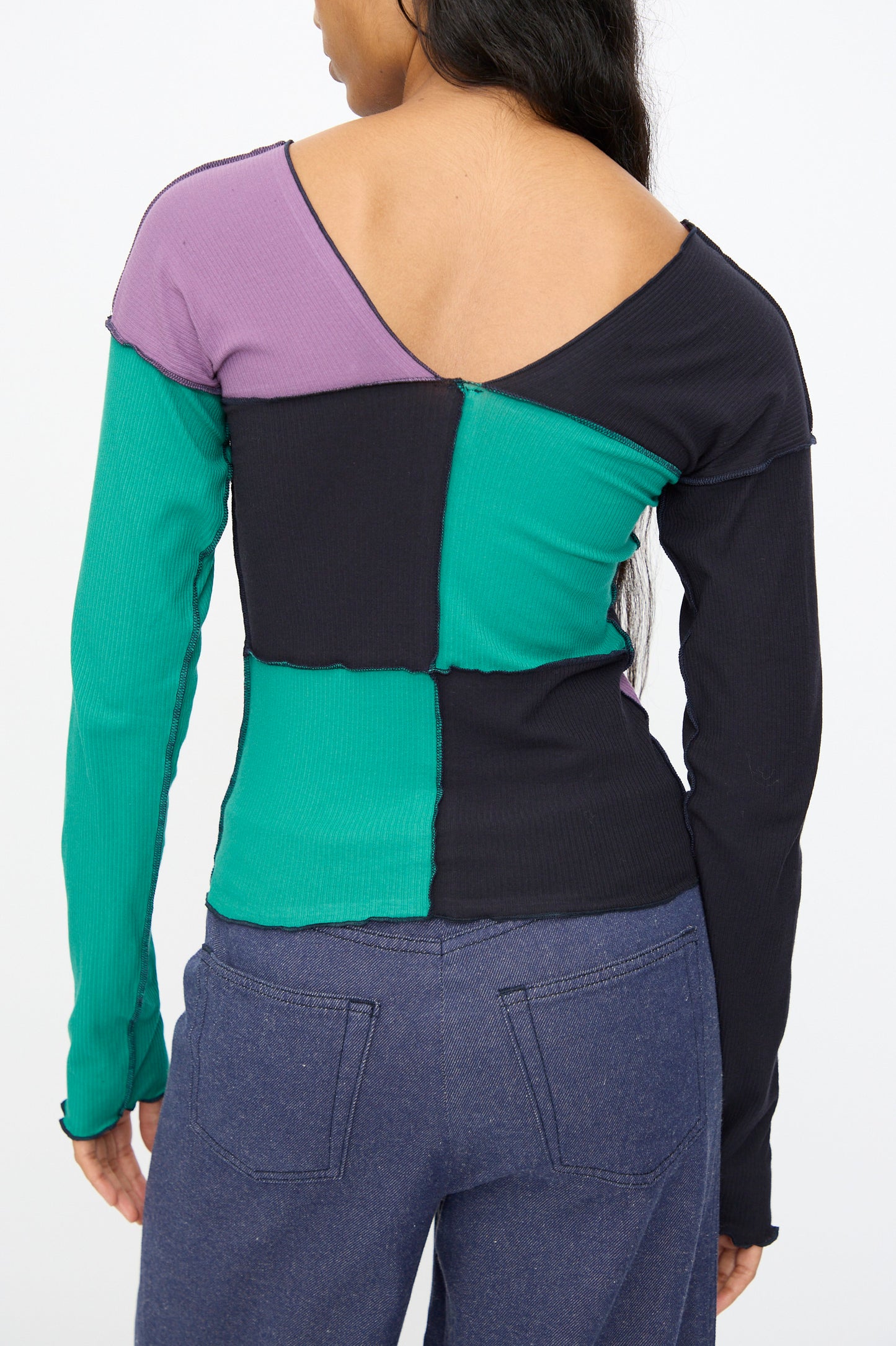 A person shown from the back wears Baserange's Teak Long Sleeve Tee in green, navy, and purple organic cotton, paired with blue jeans.