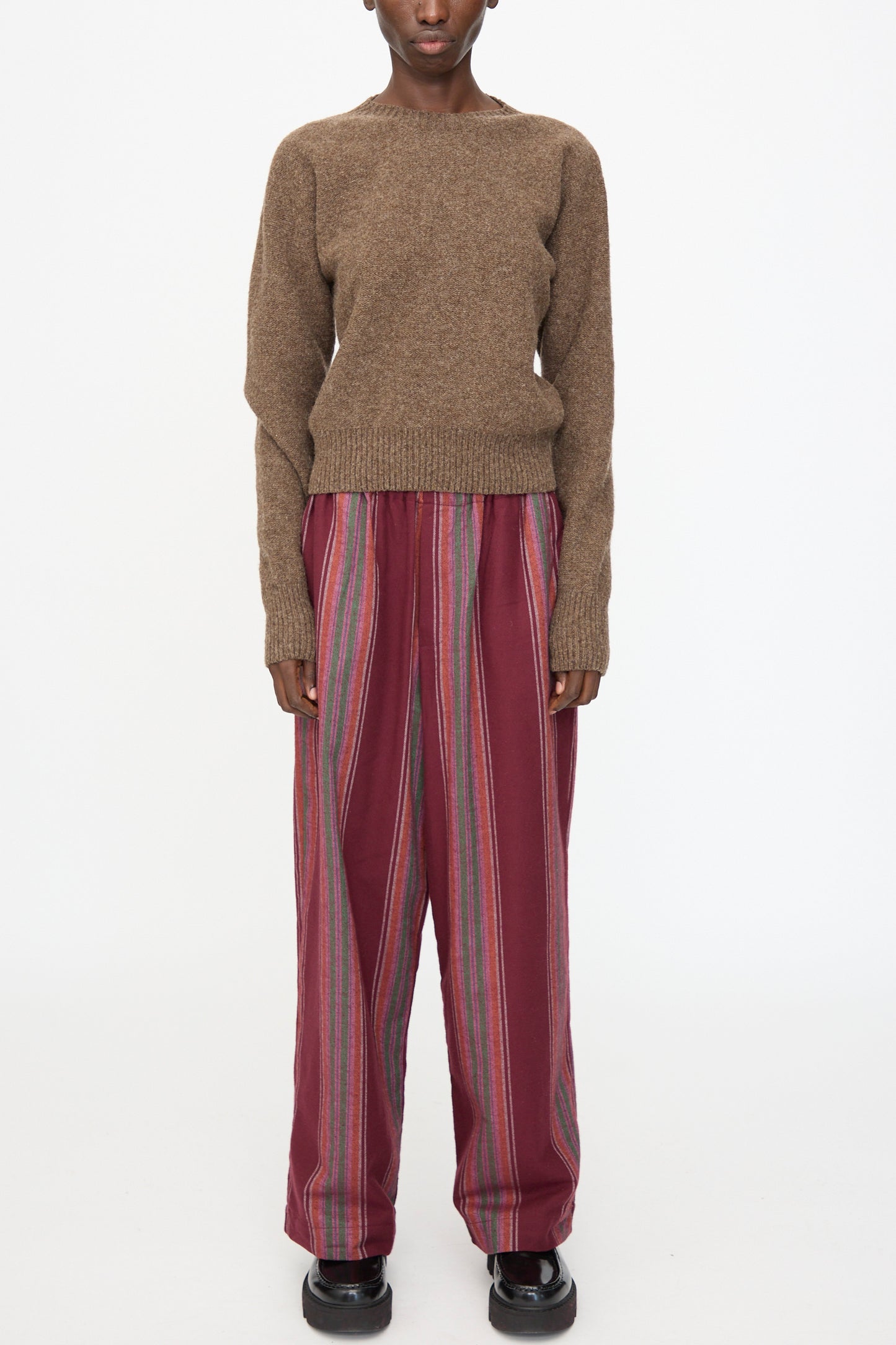 A model wears the Baserange Tier Sweater in Burel Brown, paired with loose maroon striped pants, standing against a plain white background.