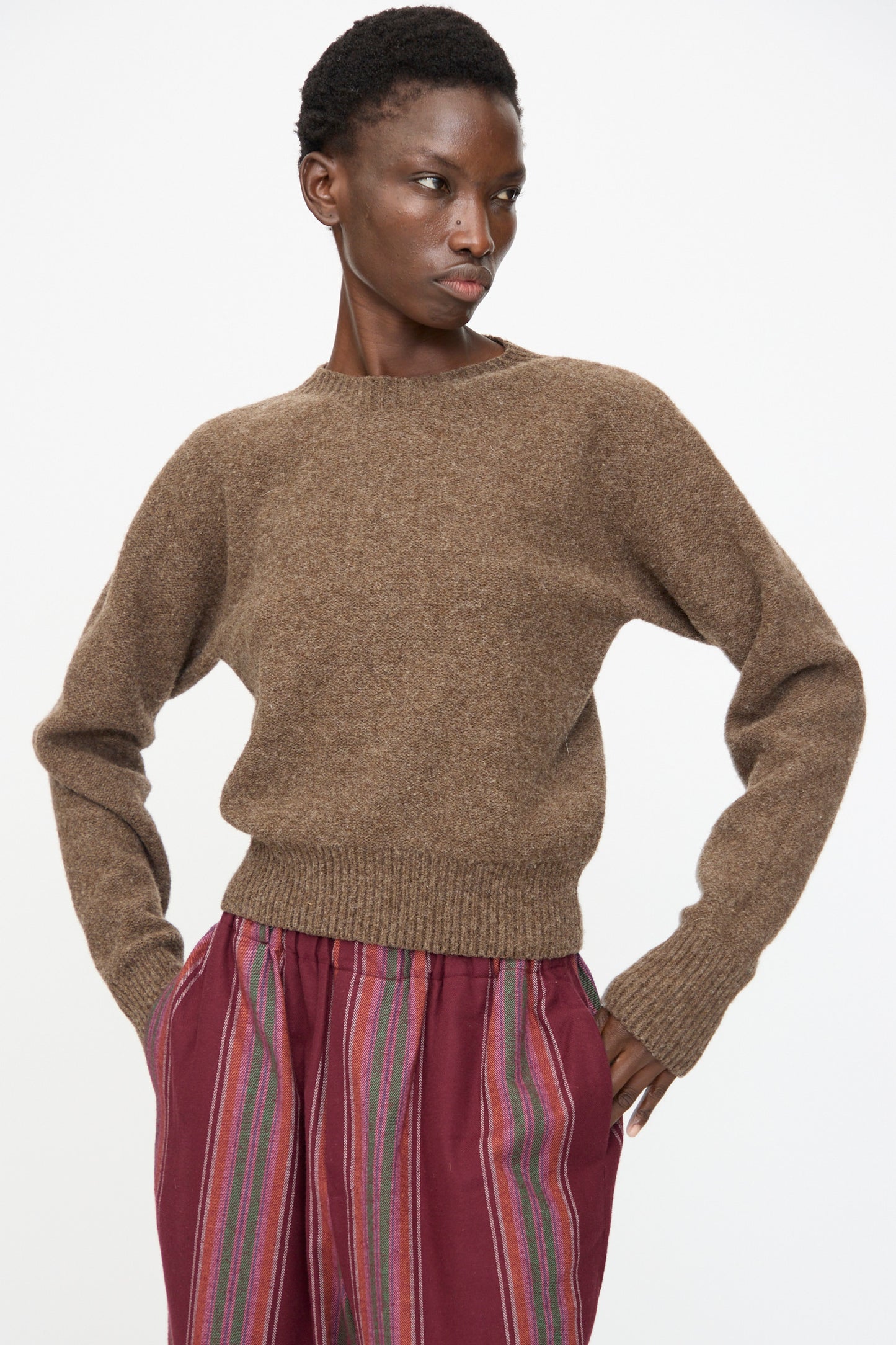 A person stands with hands in pockets, wearing the Baserange Tier Sweater in Burel Brown against a plain white background.