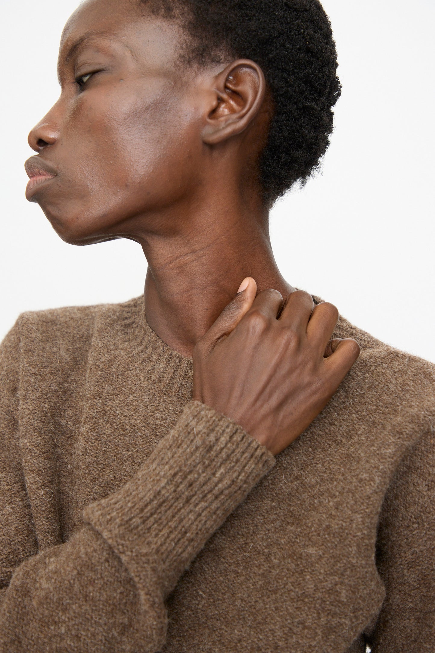 A person wearing the Baserange Tier Sweater in Burel Brown looks to the side and touches their collarbone with one hand against a plain white background.