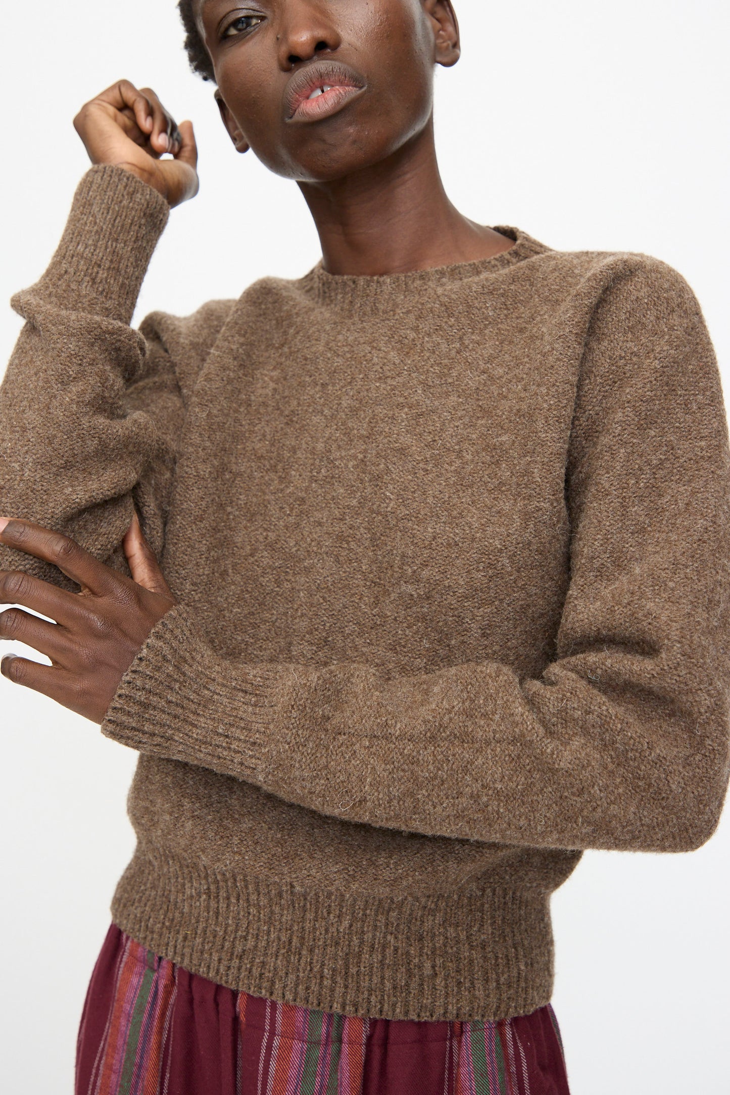 A person wearing the Baserange Tier Sweater in Burel Brown and a red skirt with vertical stripes poses against a plain white background.
