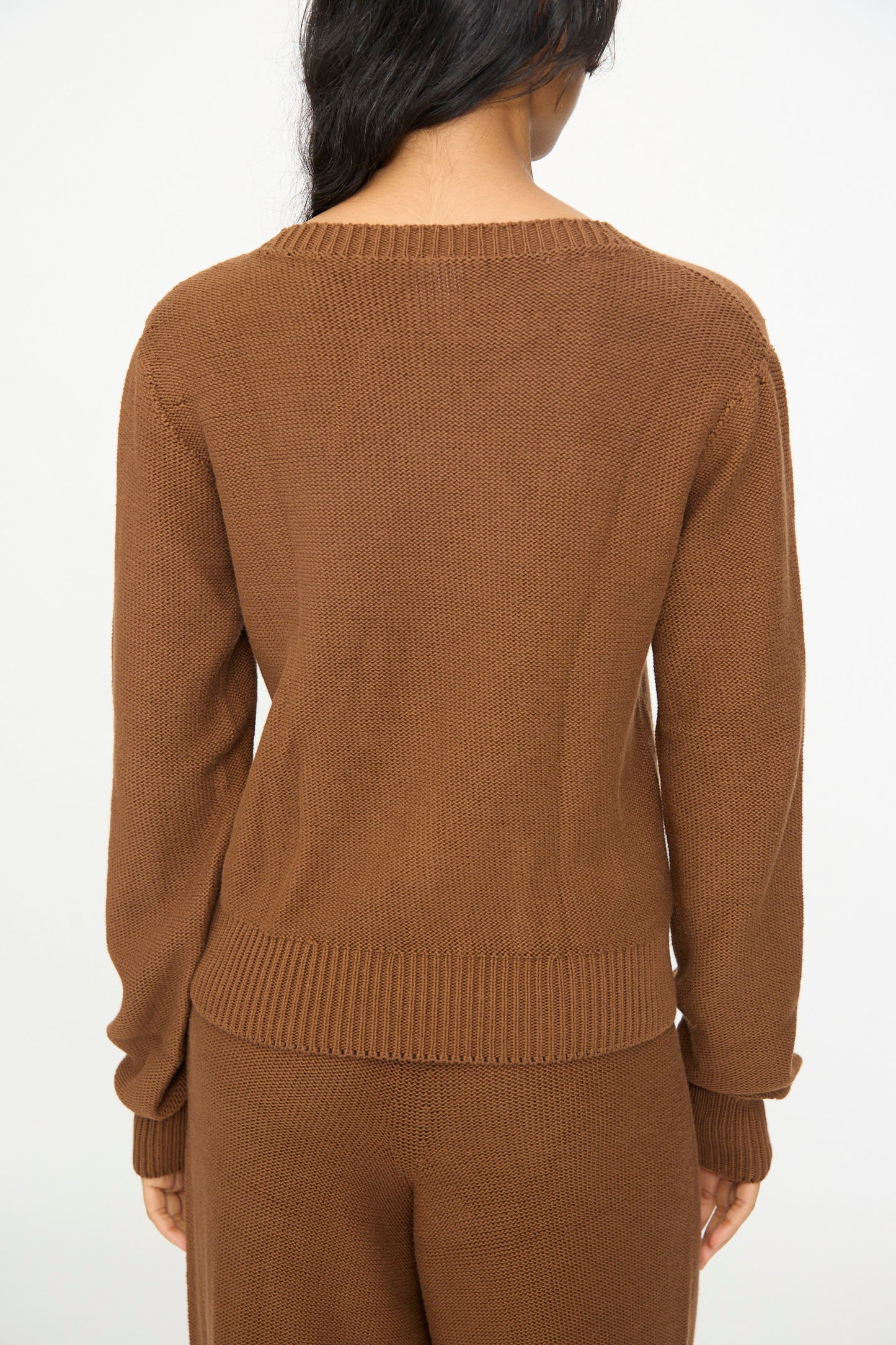 A person with long dark hair is seen from the back against a plain background, wearing the Baserange Tier Sweater in Nocciola Brown.