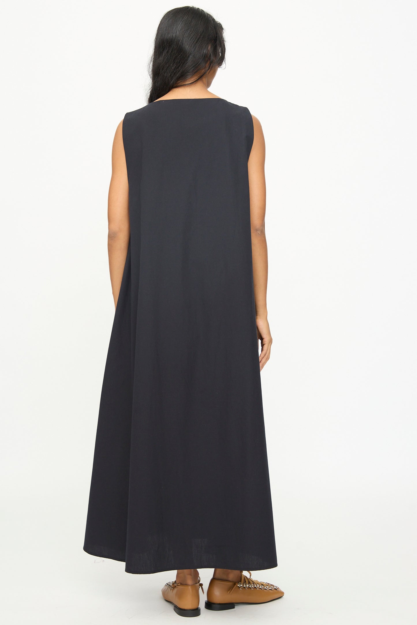 A person wearing the Baserange Tres Dress in Black—a sleeveless, relaxed-fit, ankle-length dress—stands with their back to the camera against a plain white background. The look is completed with brown shoes for an understated effect.