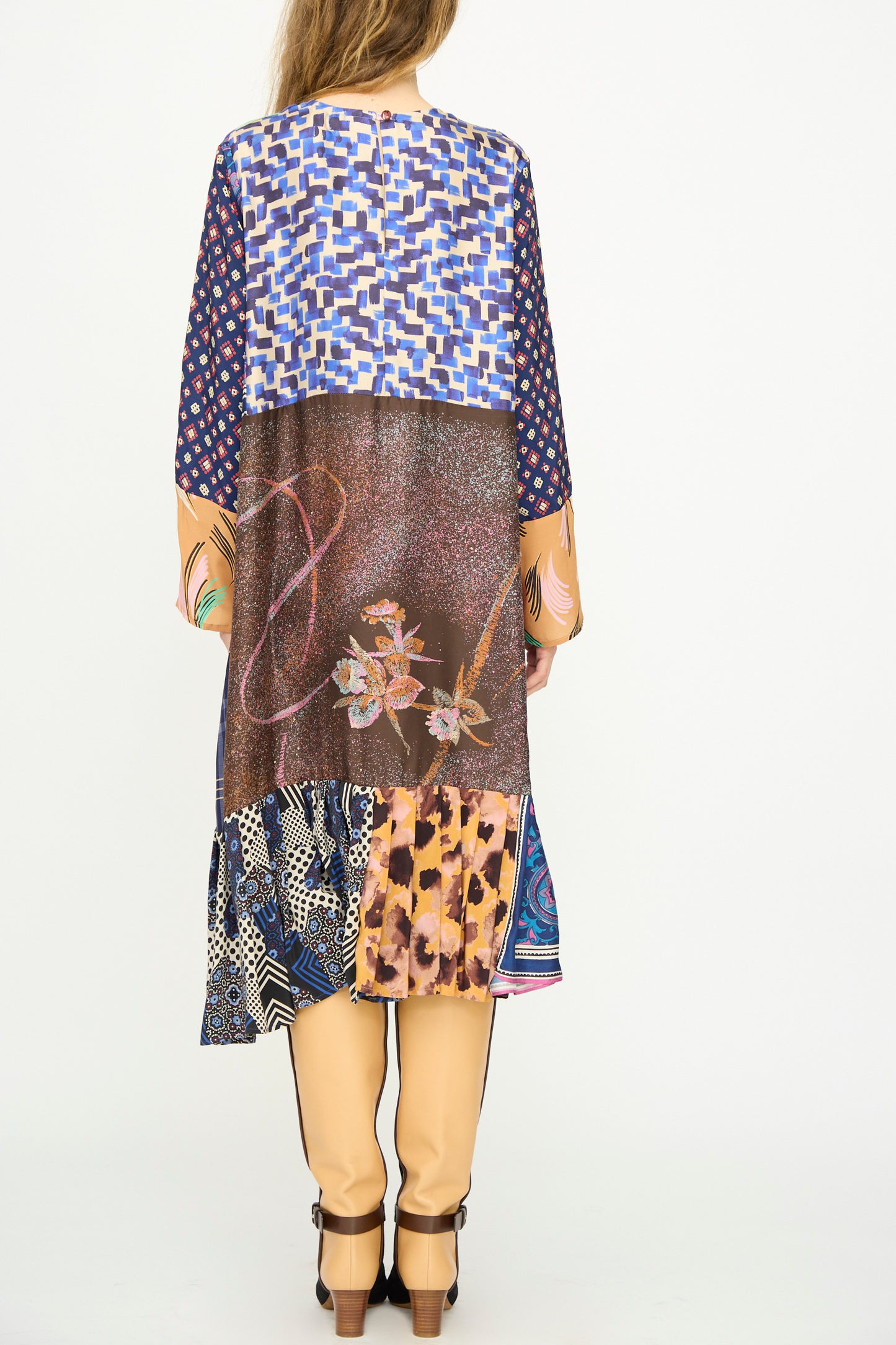 A person faces away, wearing the Bettina Bakdal Hannah Silk Dress in Ricarda Meltzer—a patchwork of vintage silk scarves—paired with light tan pants and heeled shoes.