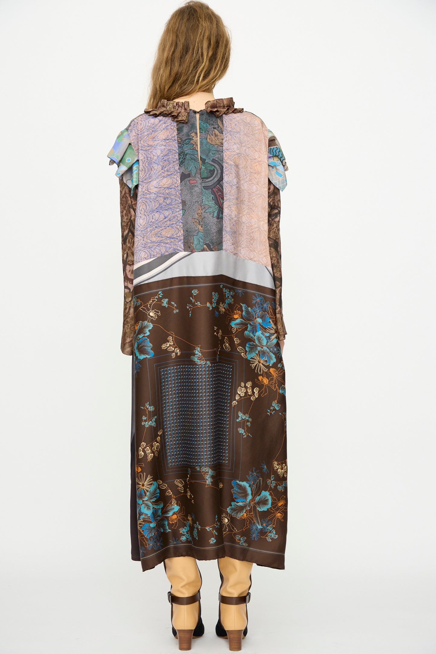 A person stands facing away, wearing the Bettina Bakdal Ruffled Collar and Shoulder Vintage Scarves Dress in Margarete Dambeck, featuring floral and geometric patterns, paired with tan pants and black shoes.