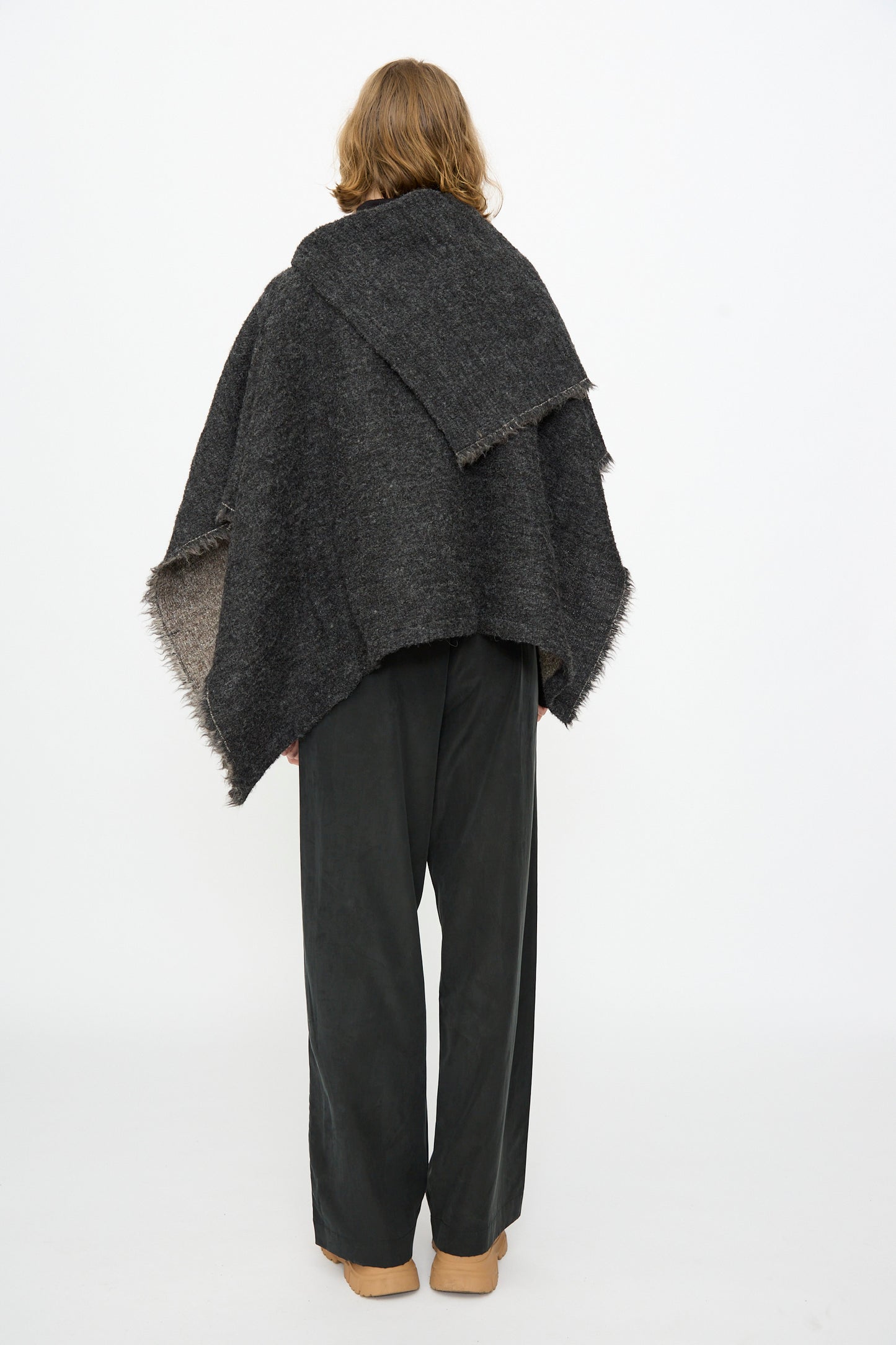A person with shoulder-length hair stands facing away against a plain white background, wearing the Black Crane Alpaca Short Poncho in Dark Grey paired with loose black pants and tan shoes.