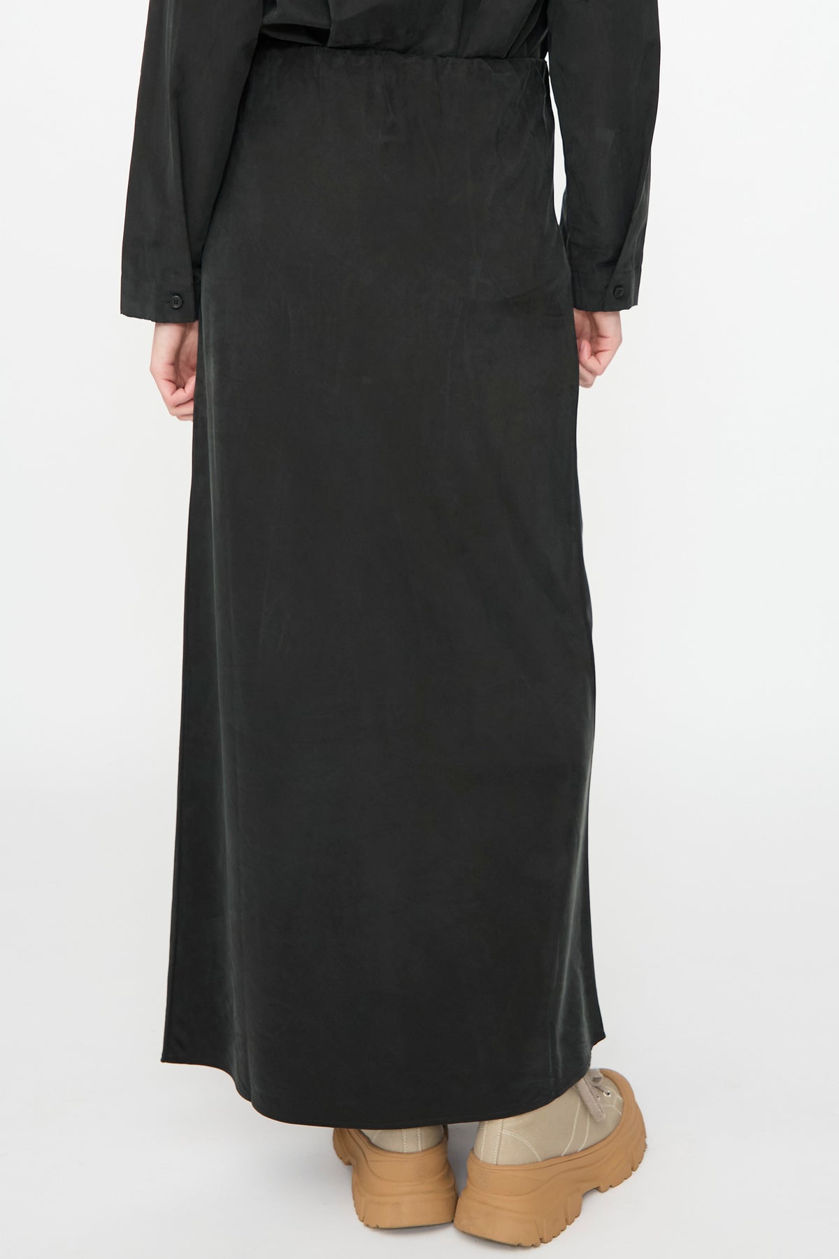 A person is shown from the back against a plain white background, wearing the Black Crane Bias Skirt in Black—a long maxi skirt crafted from a soft cotton cupro blend with an elastic waistband for added comfort—paired with tan sneakers.