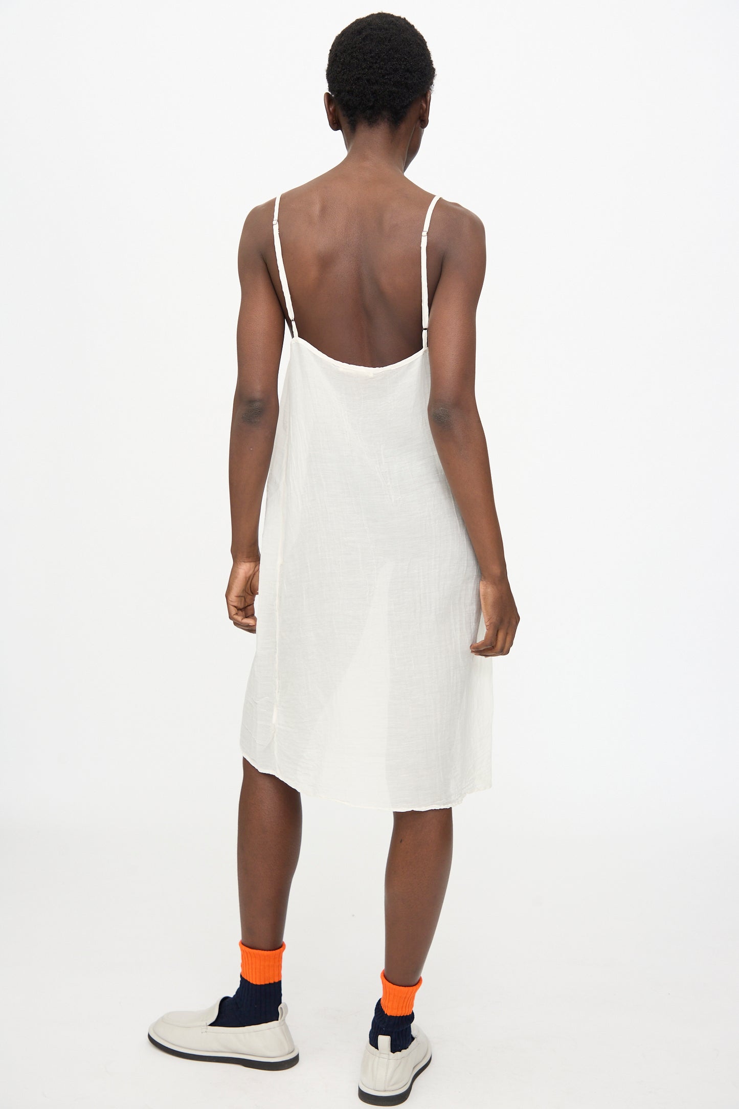 A person stands with their back to the camera wearing the Black Crane Cami Dress in White, paired with white sneakers and orange socks, against a plain white background.