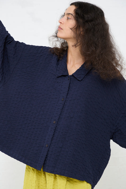 A person with long curly hair models an oversized fit, textured navy blue Black Crane Cotton Seersucker Chelsea Collar Shirt while gazing to the side.