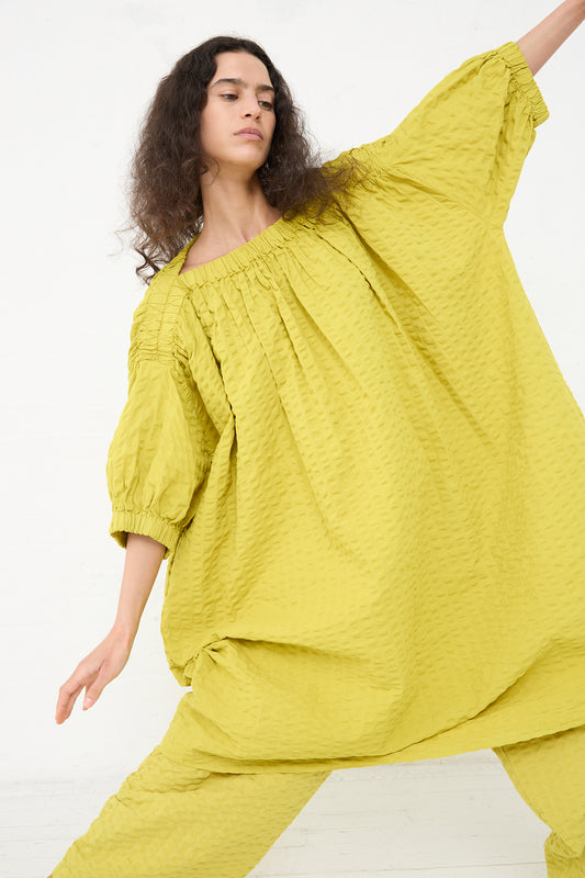 A woman is posing in a spacious room, wearing a loose-fitting, yellow Black Crane cotton seersucker sack dress with a textured pattern. She has curly hair and is extending one arm out to the side.