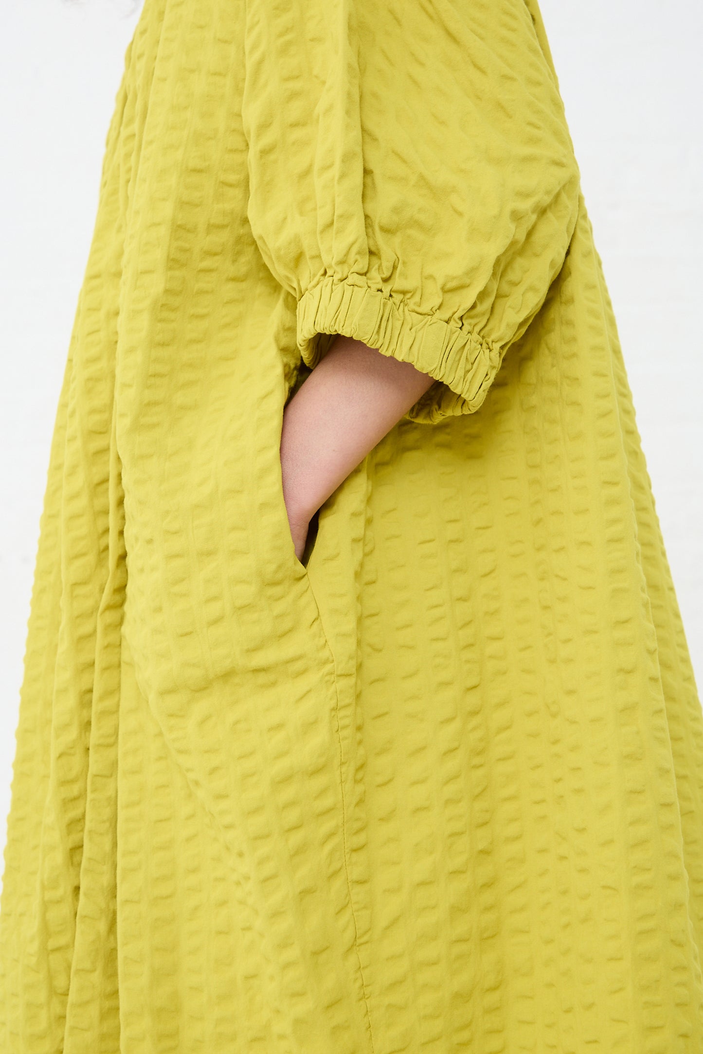 A hand protrudes from a slit in a textured yellow Cotton Seersucker Sack Dress in Turmeric by Black Crane.