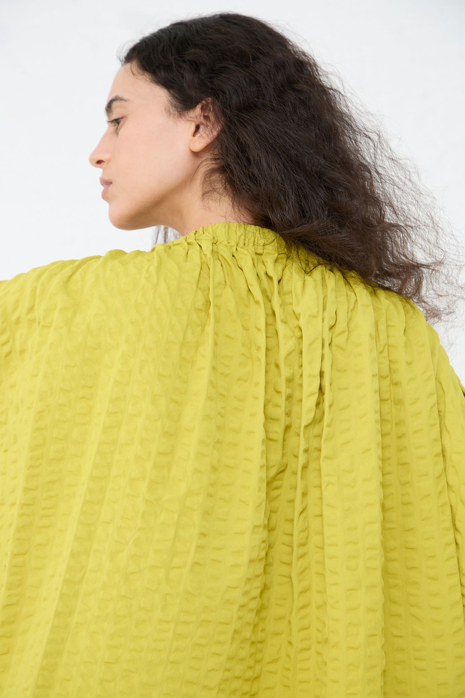 A woman in a Black Crane cotton seersucker sack dress in turmeric is seen from a side angle against a plain background, with her face turned away from the camera.