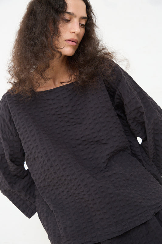 A woman with curly hair wearing a textured Black Crane Cotton Seersucker Square Neck Top in Ink Black gazes downward to her left with a contemplative expression.