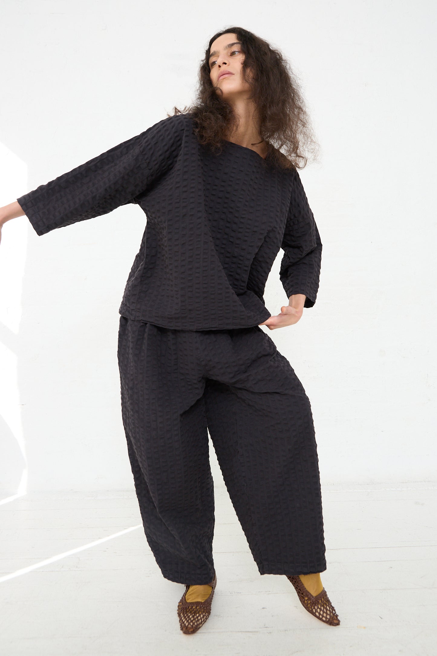 A person standing in a relaxed pose wearing a textured dark Black Crane Cotton Seersucker Square Neck Top in Ink Black outfit with matching pants, complemented by patterned flat shoes.