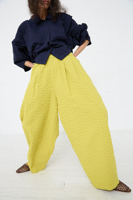 A person standing against a white background wearing a navy blue textured blouse tucked into high-waisted, pleated Cotton Seersucker Wide Pant in Turmeric from Black Crane and dark woven shoes.
