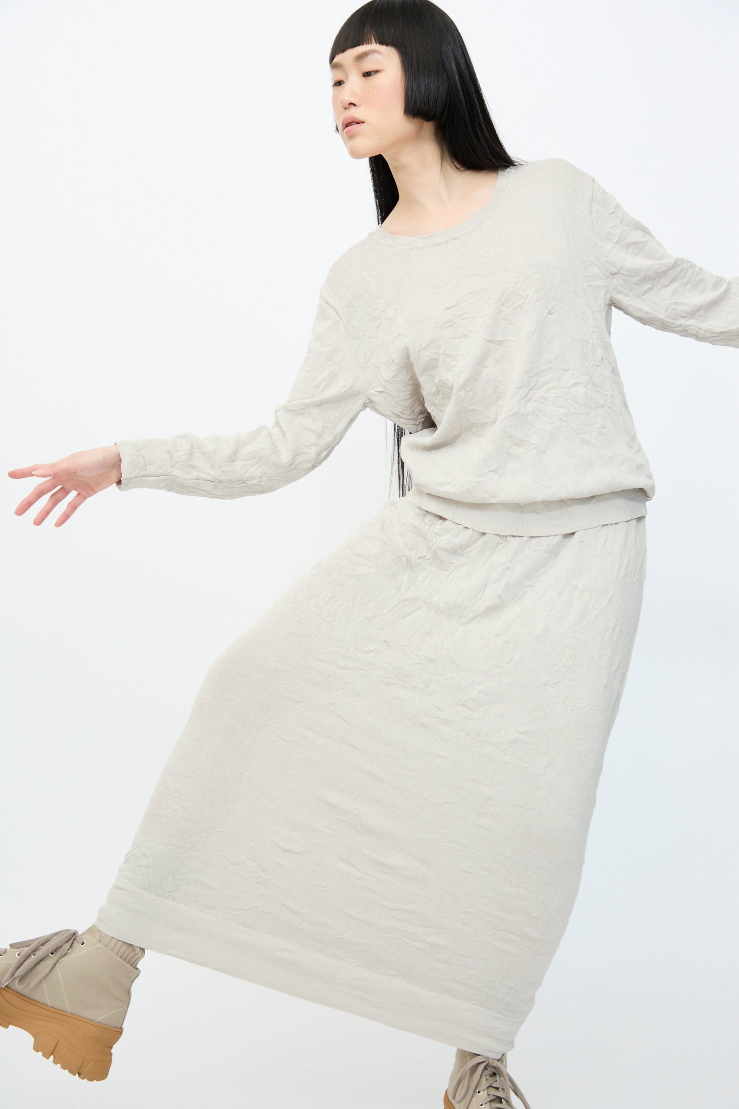 Wearing the Crinkled Knit Skirt in Platinum by Black Crane, a person poses against a plain backdrop. Their outfit is made from recycled polyester, complemented by matching beige shoes.