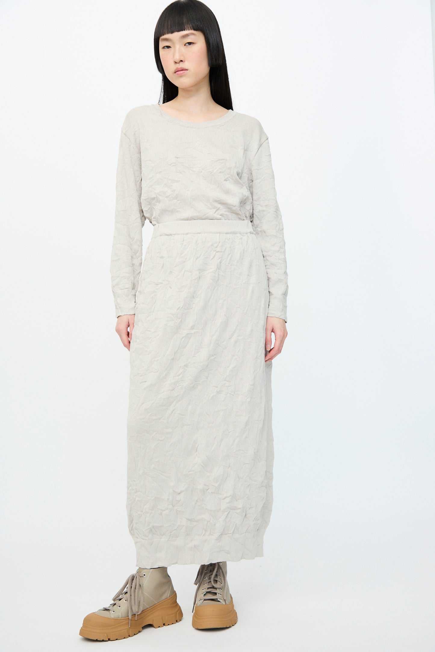 A person with long hair in a Crinkled Knit Skirt in platinum by Black Crane and tan platform boots made from recycled polyester stands against a plain white background.