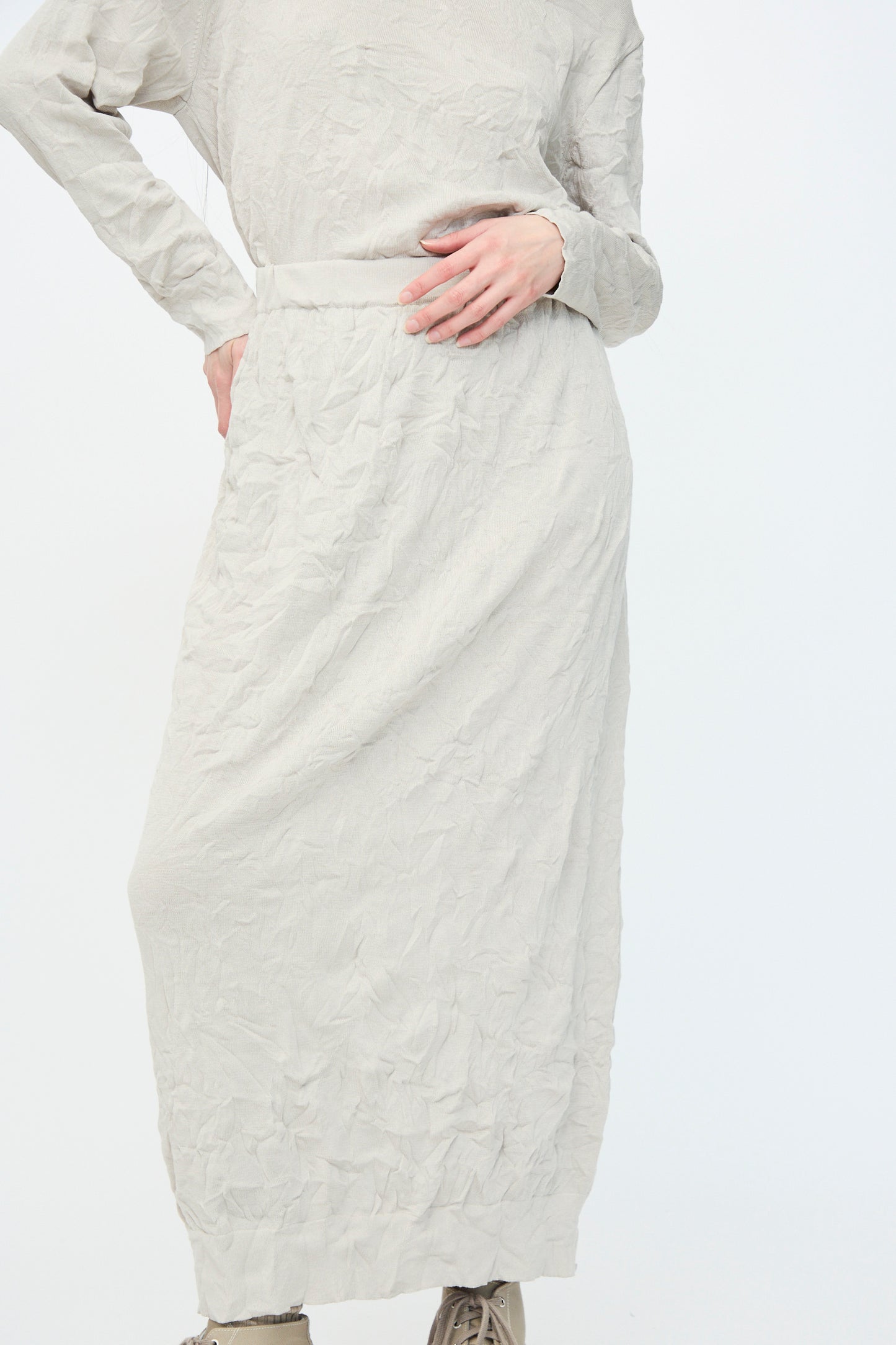 A person dressed in Black Crane's Crinkled Knit Skirt in Platinum made from recycled polyester, with their hand on their waist against a white backdrop.