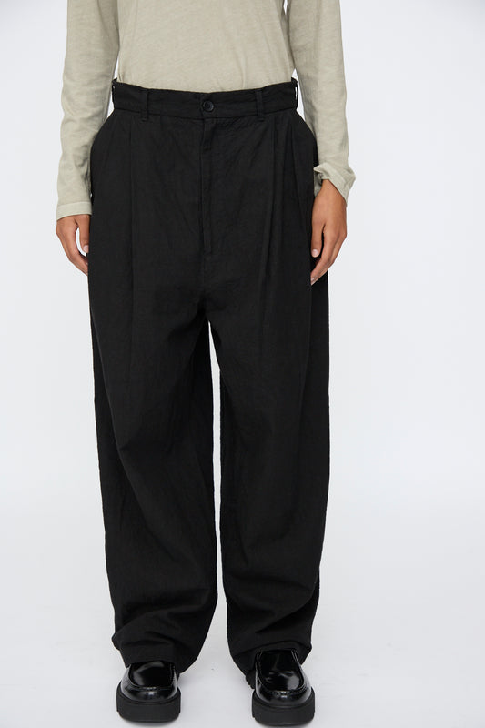 A person wearing Black Crane Crinkled Trousers in Black, a light beige long-sleeve shirt, and black shoes stands against a plain white background.