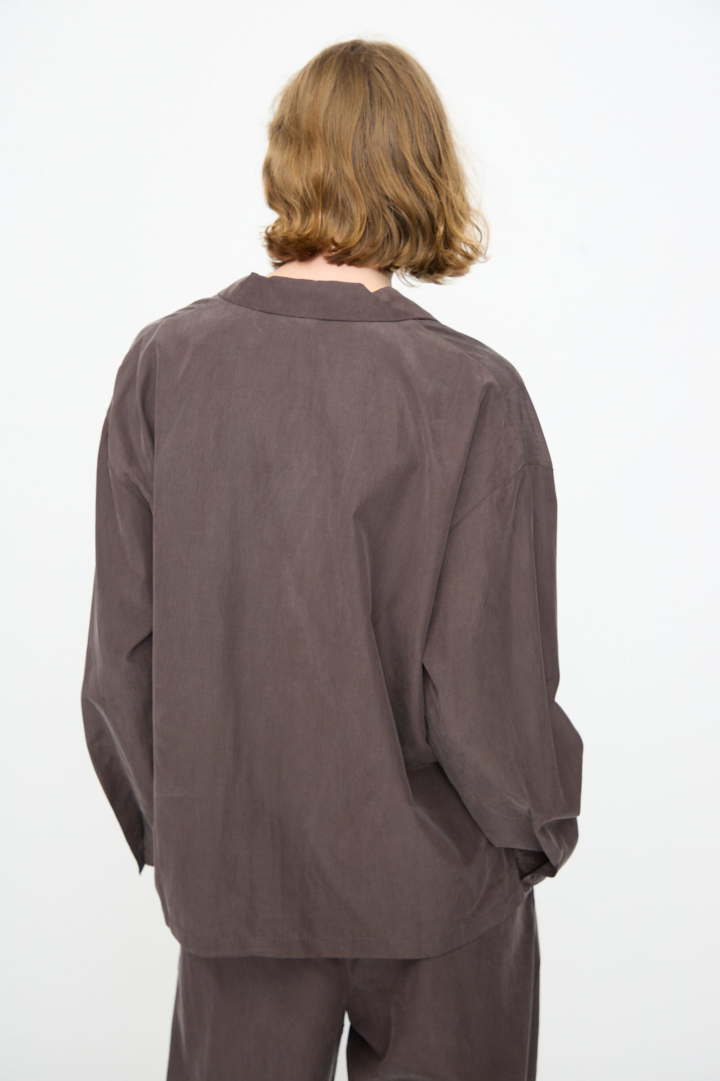 A person with shoulder-length light brown hair is seen from the back against a plain white background, wearing Black Crane’s Cropped Shirt in Dark Plum—a loose-fitting, boxy long-sleeved top crafted from a soft cotton cupro blend.