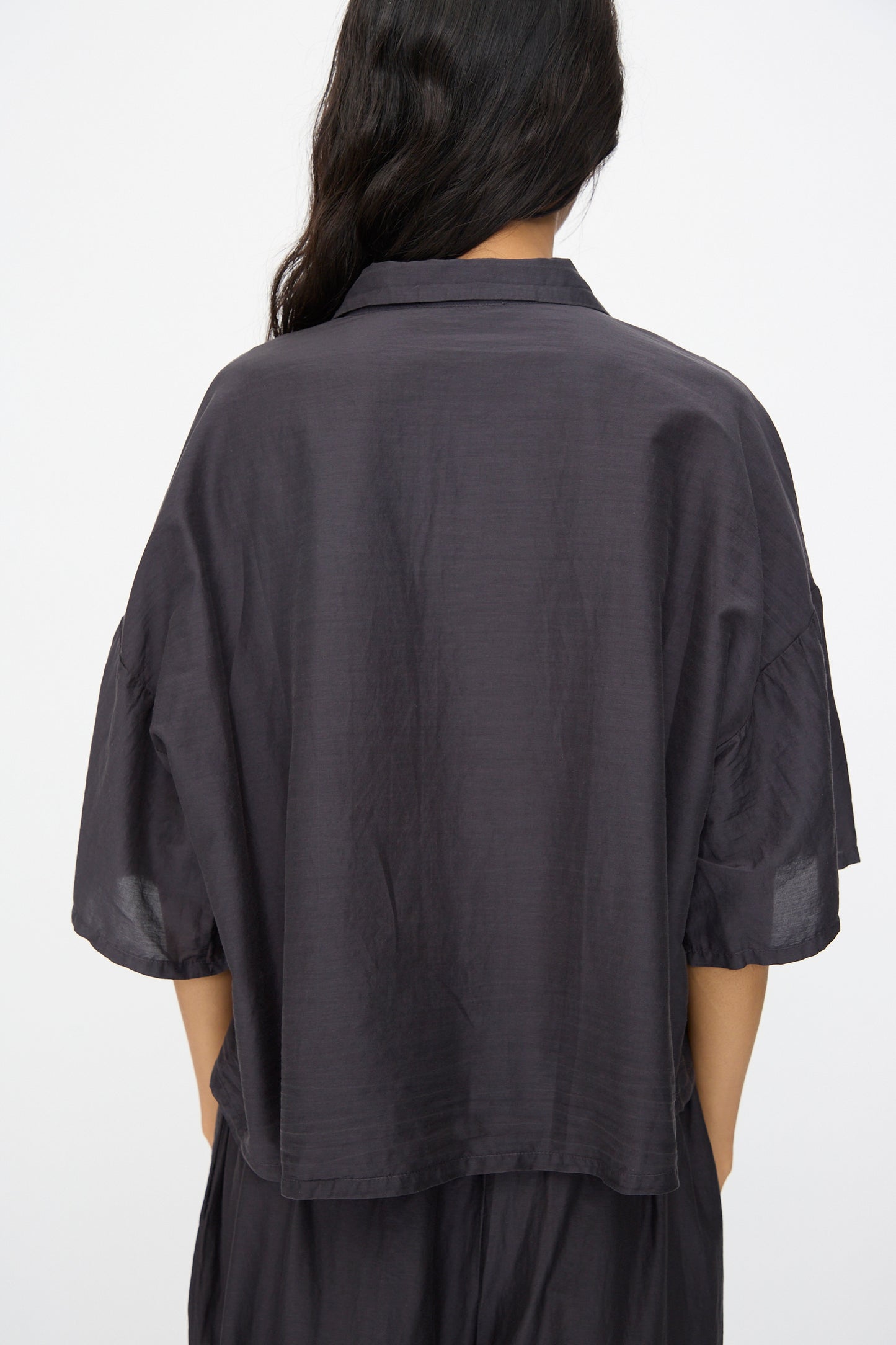 A person with long dark hair is seen from behind wearing the Black Crane Cropped Shirt in Faded Black, a loose, boxy-fit short sleeve top made from a soft cotton silk blend, set against a plain background.