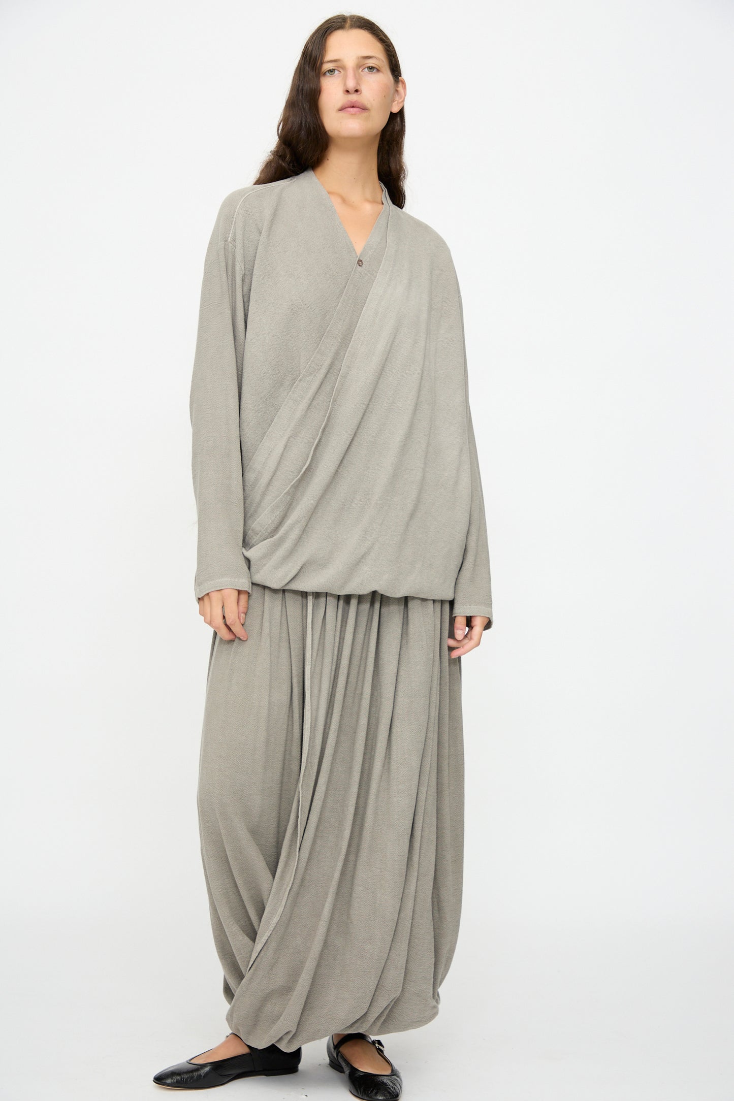 A person stands against a plain background wearing Black Crane's Double Layered Drapy Skirt in Dark Stone—a loose, cotton rayon blend midi with long sleeves—paired with black flat shoes.