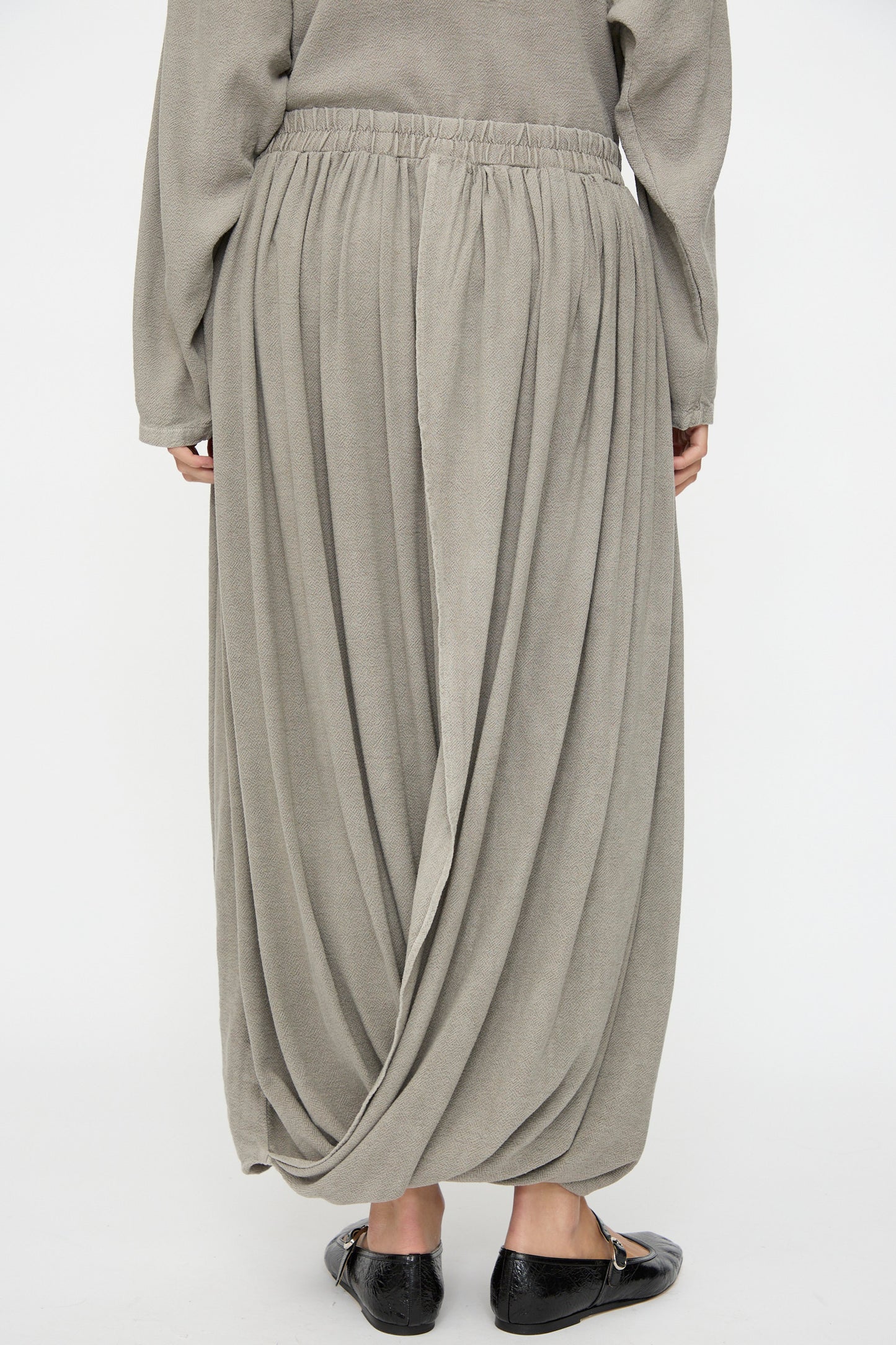 A person is seen from the back wearing the Black Crane Double Layered Drapy Skirt in Dark Stone, a light grey cotton-rayon midi with an elastic waistband, paired with black flat shoes.