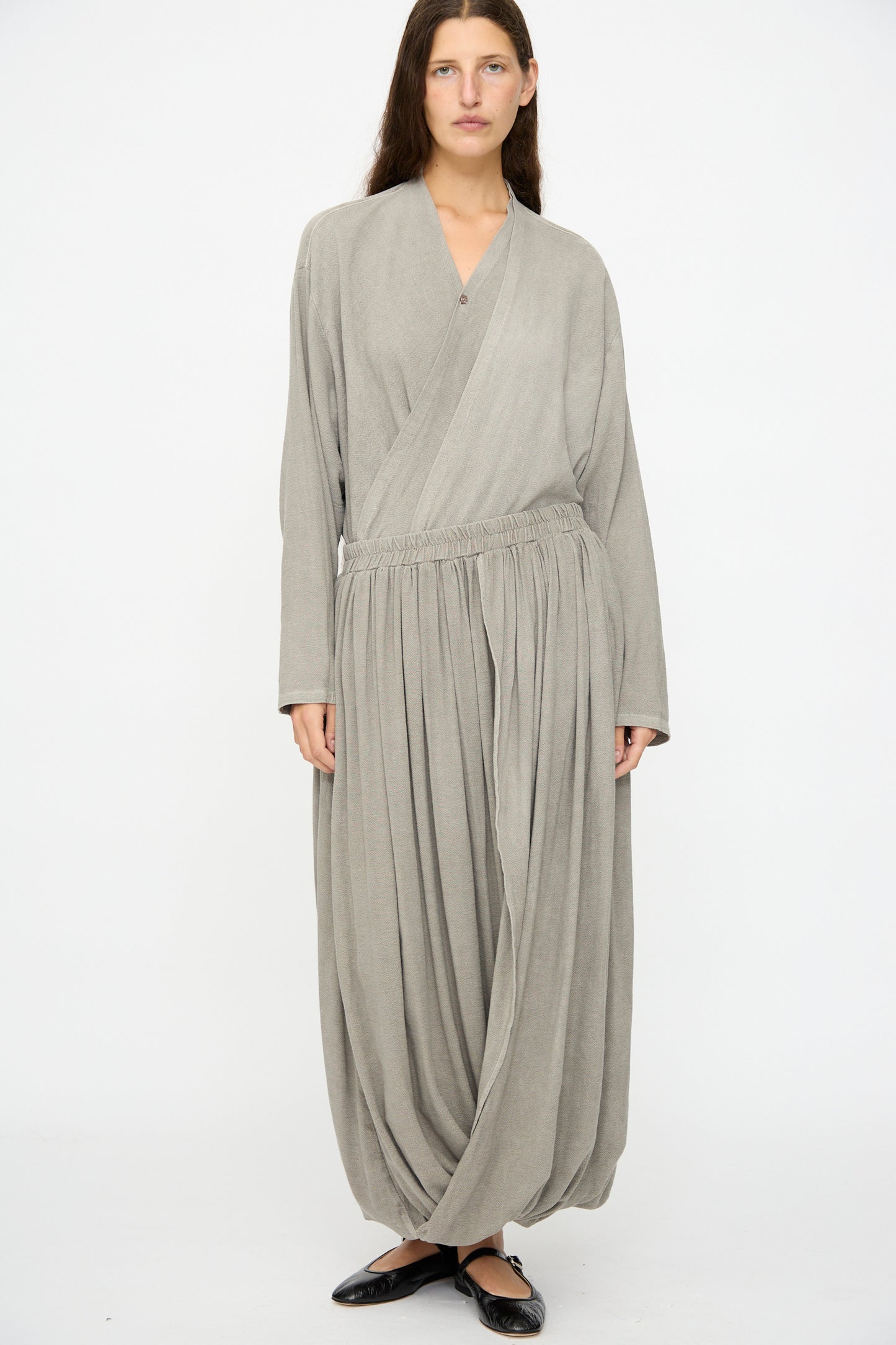 A person stands against a plain background wearing a loose, light gray wrap top paired with Black Crane's Double Layered Drapy Skirt in Dark Stone, featuring an elastic waistband and wide drape, finished with black flat shoes.