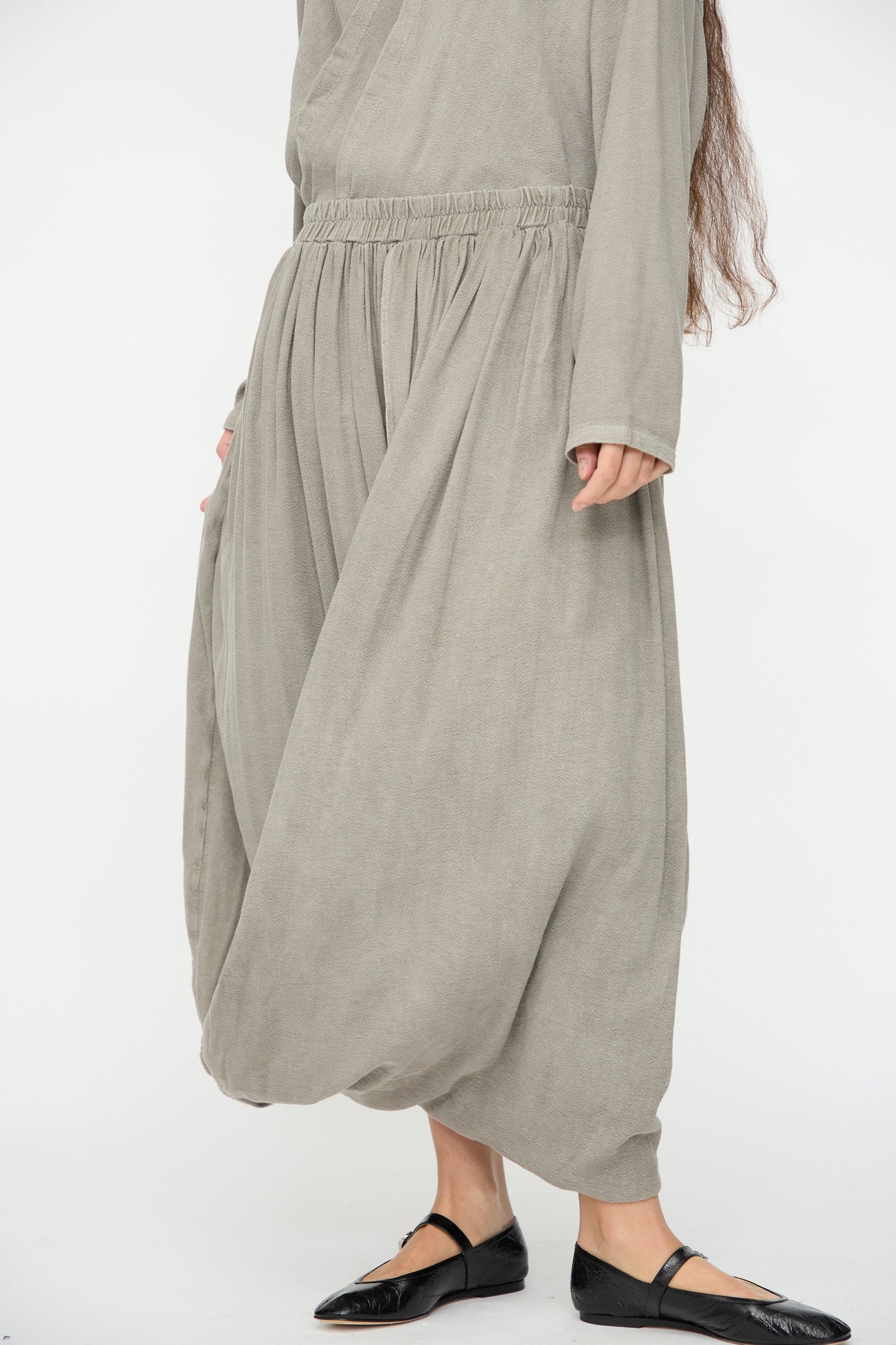 A person wearing Black Crane's Double Layered Drapy Skirt in Dark Stone with a loose, light gray long-sleeved top and black pointed flats stands against a plain white background.