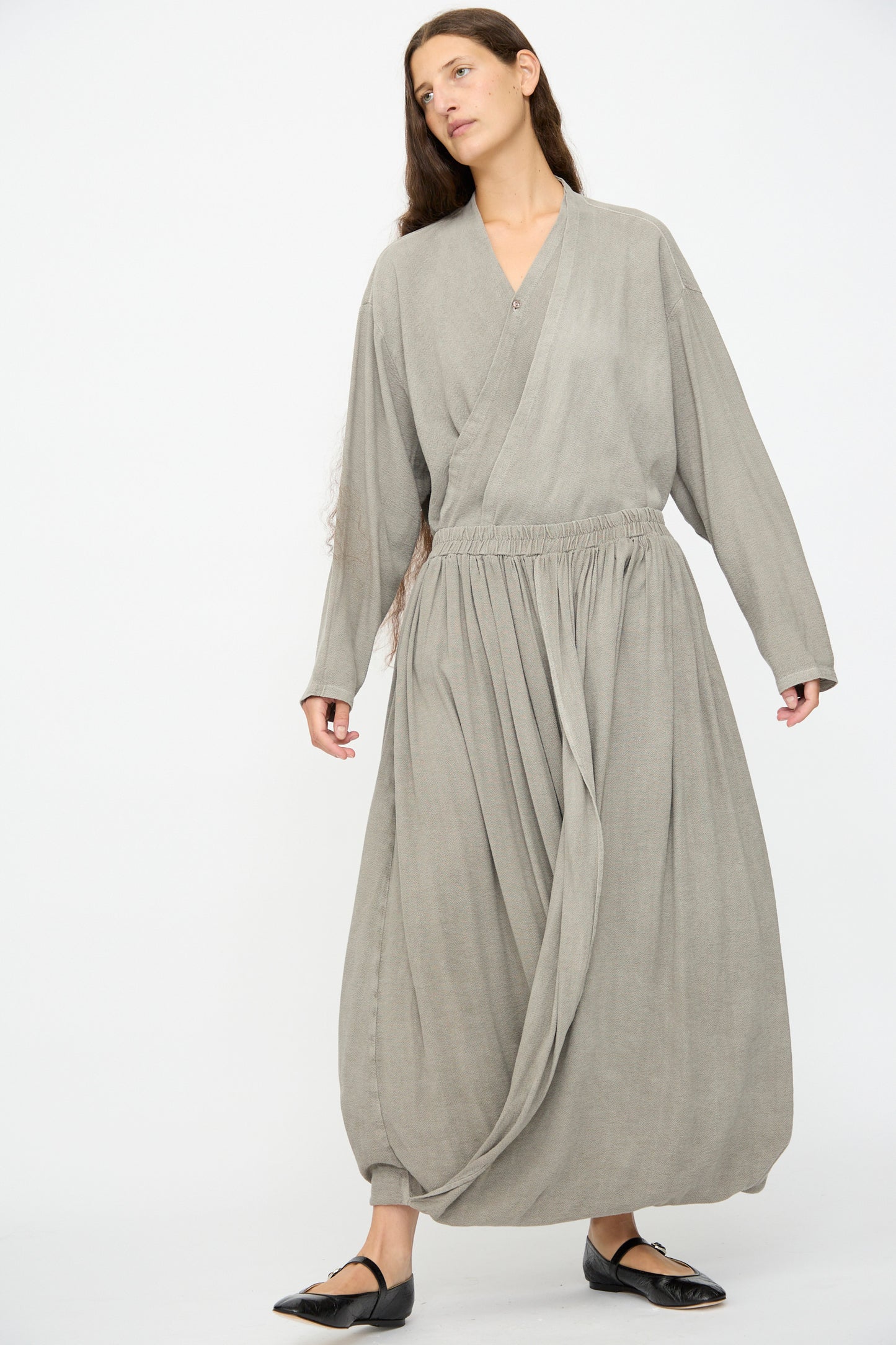 A person with long hair wears the Black Crane Double Layered Drapy Skirt in Dark Stone, styled with a loose-fitting, light grey, long-sleeved wrap top. They are standing against a plain white background.